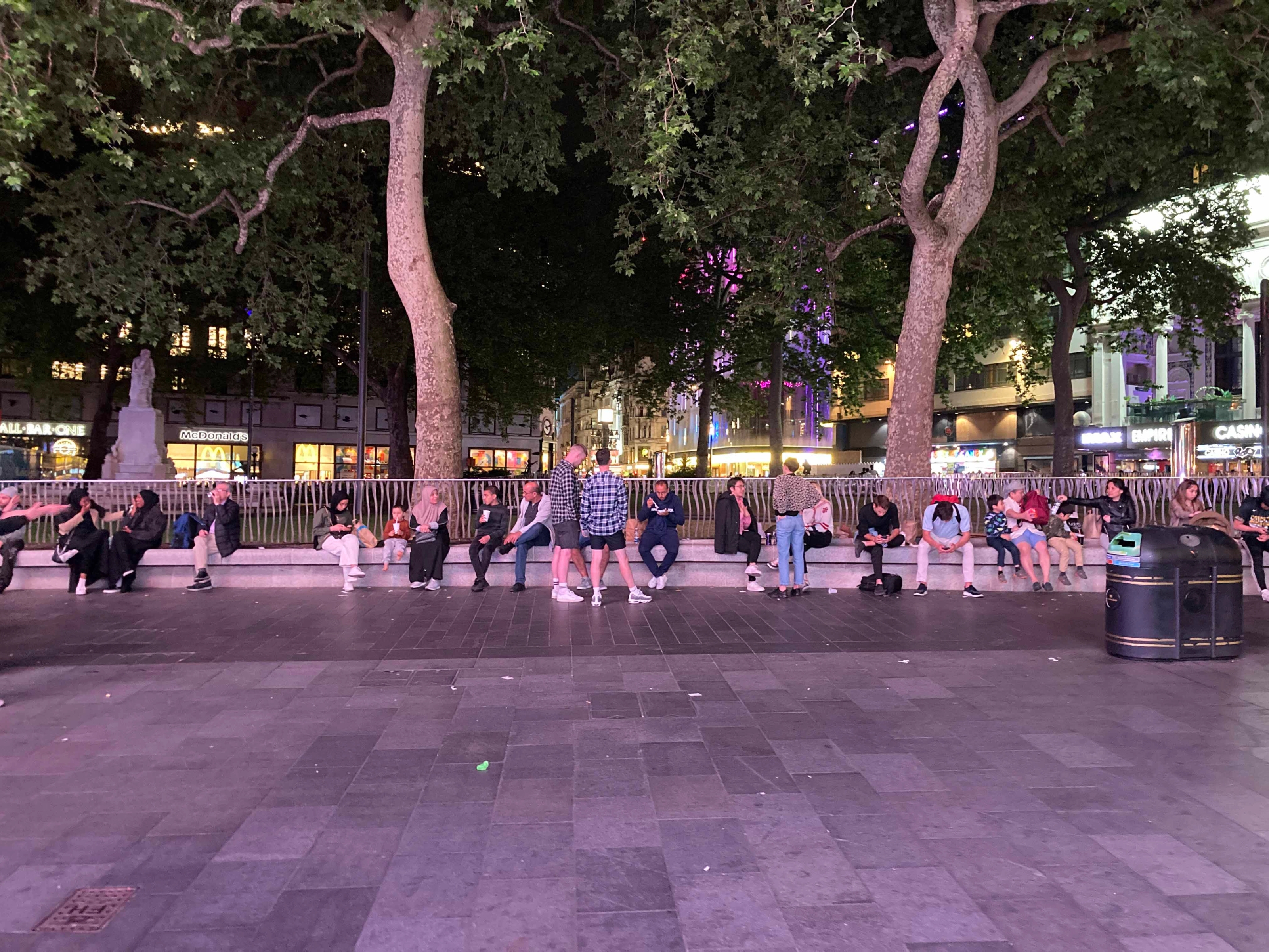 People sitting on a continuous bench around the fence of Leicester Square Gardens.