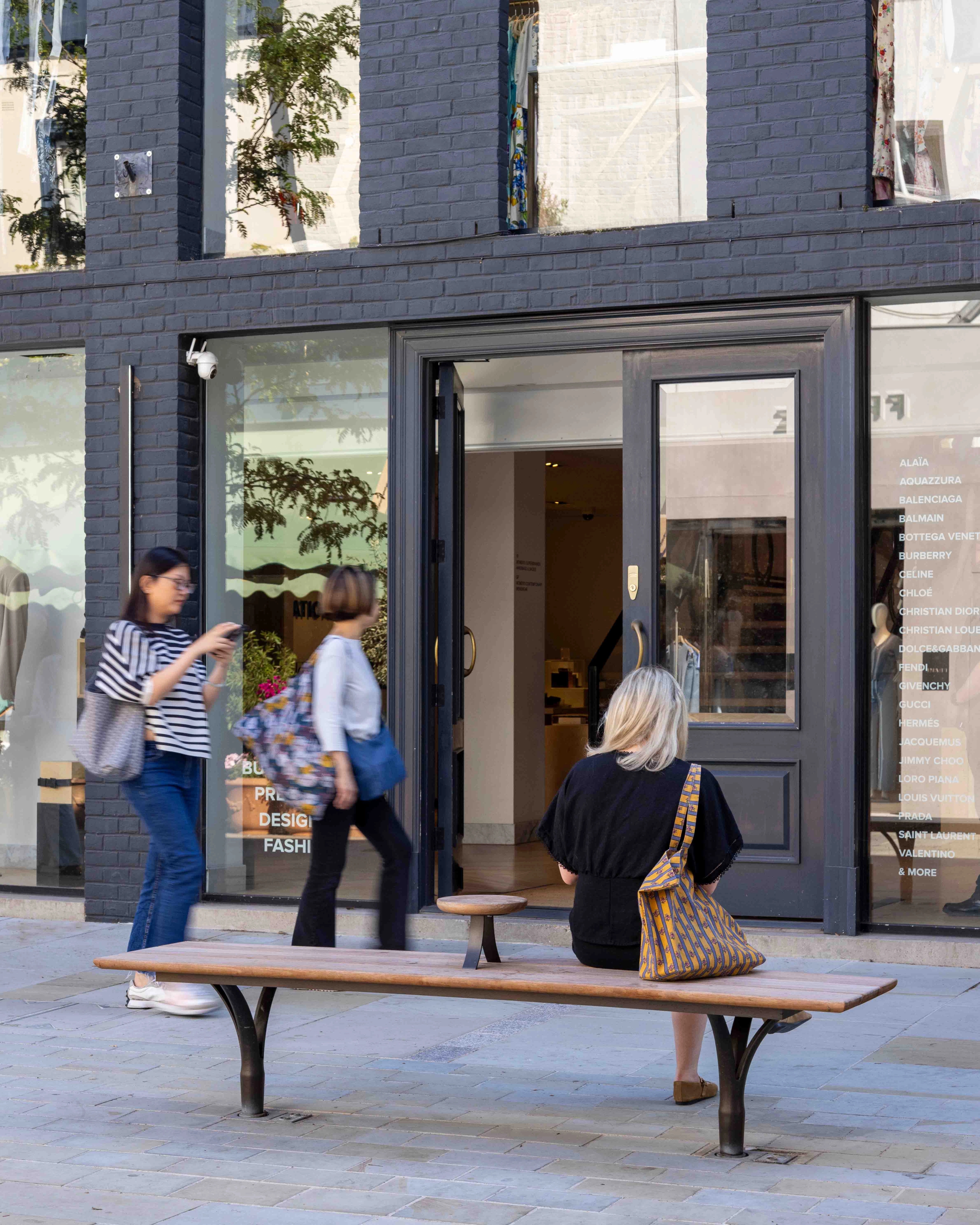 Pedestrians walking past woman sitting on bench facing away from the camera.