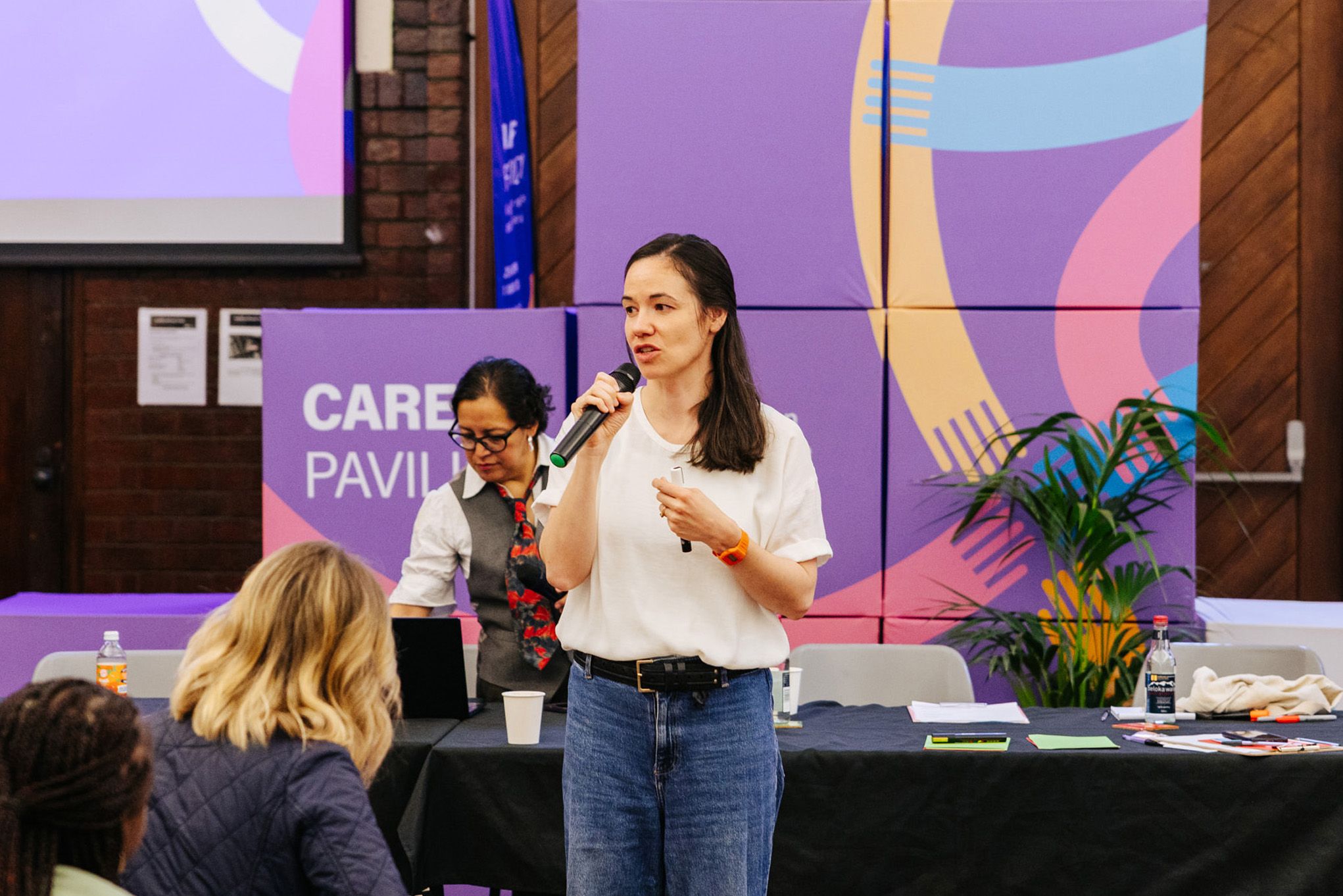 A woman in a white t-shirt and jeans speaks into a handheld microphone at an indoor event. Behind her, a colorful purple backdrop reads 'Care Pavilion', with abstract shapes in pink, blue, and orange. A second woman stands at a table in the background, and audience members are visible in the foreground.