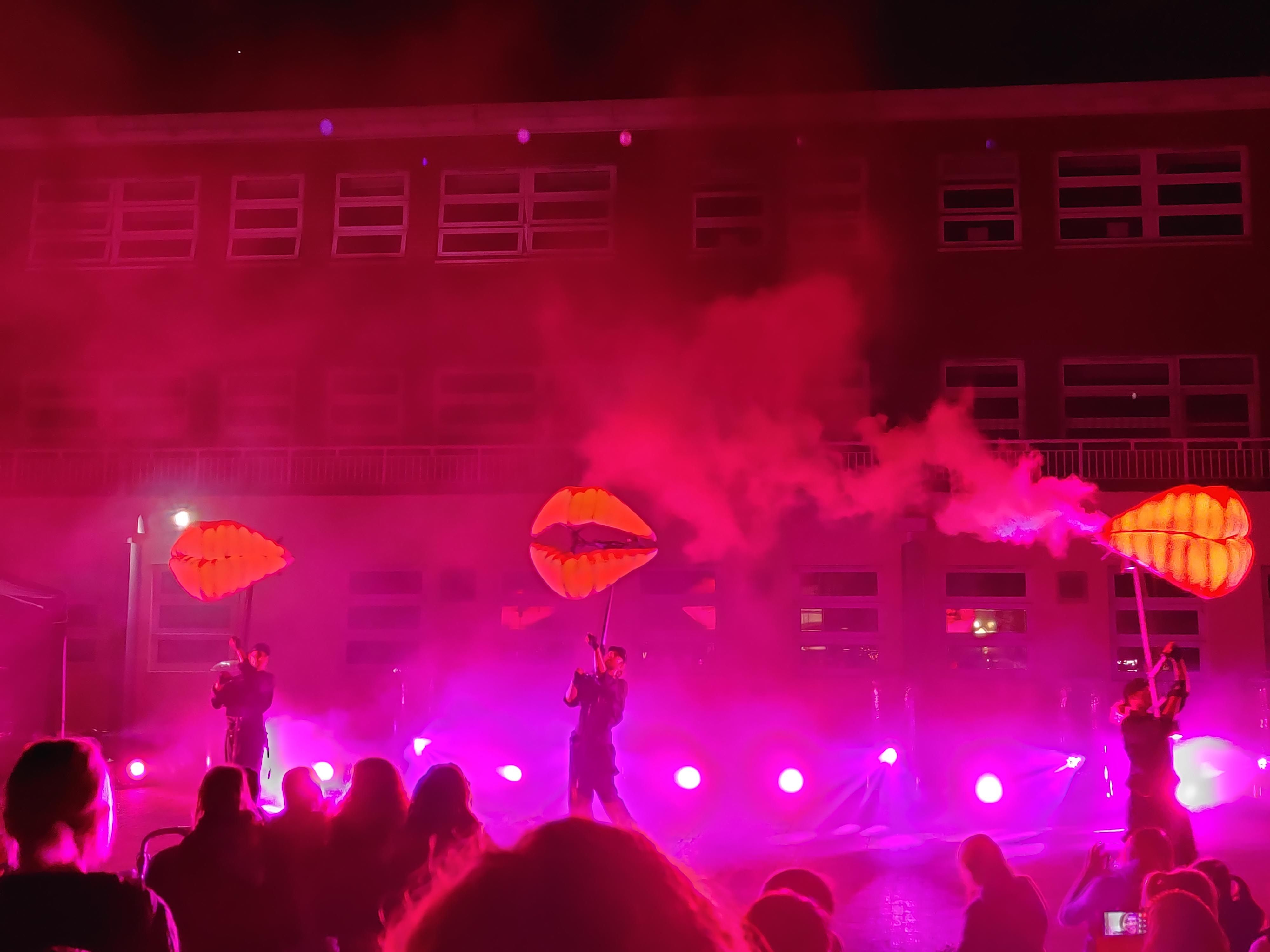 Three people on a pink lit stage infront of a crowd holding large structures with smoke.