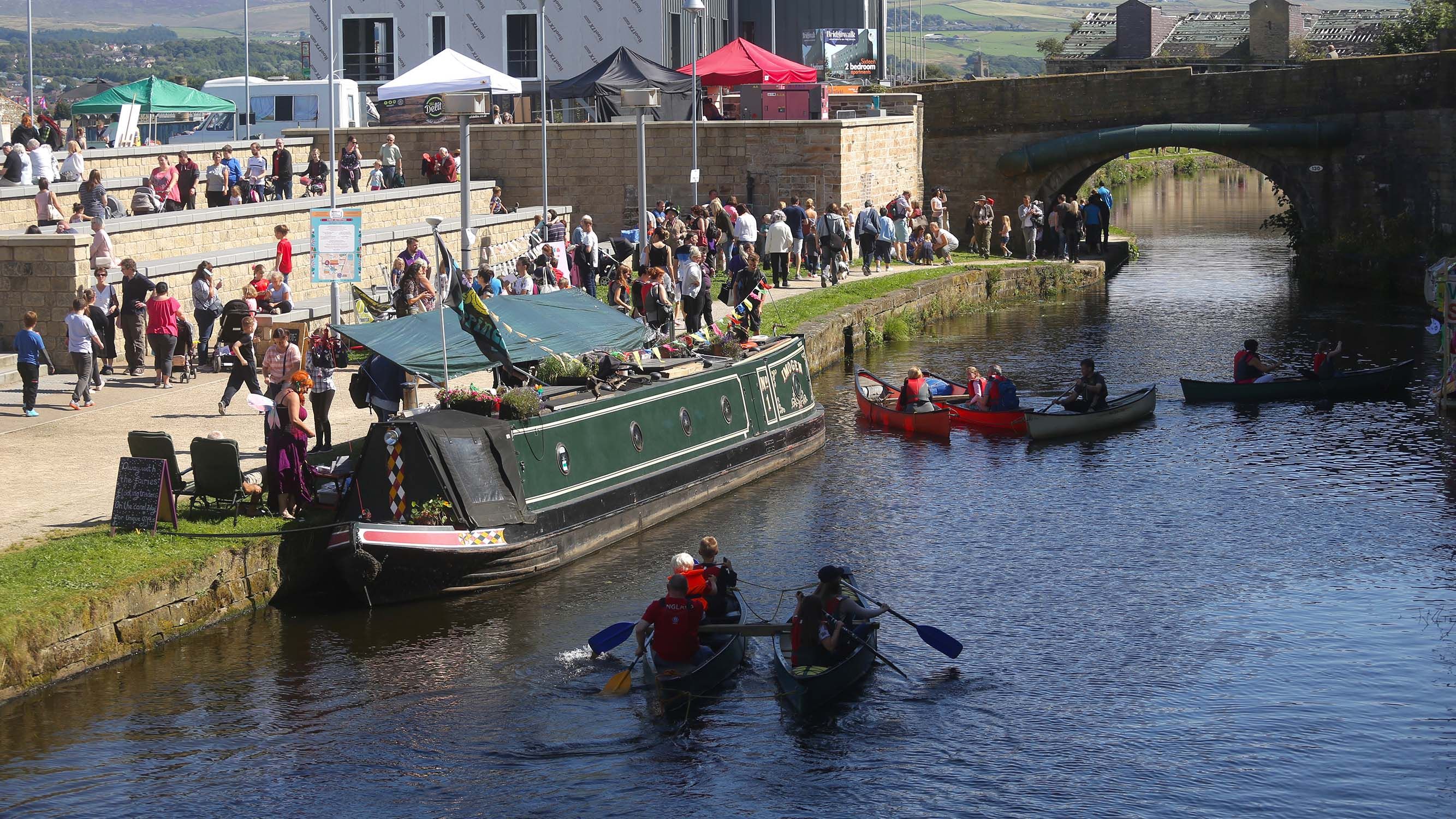 A canal on a sunny day with a boat and rowers and many pedestrians walking down.