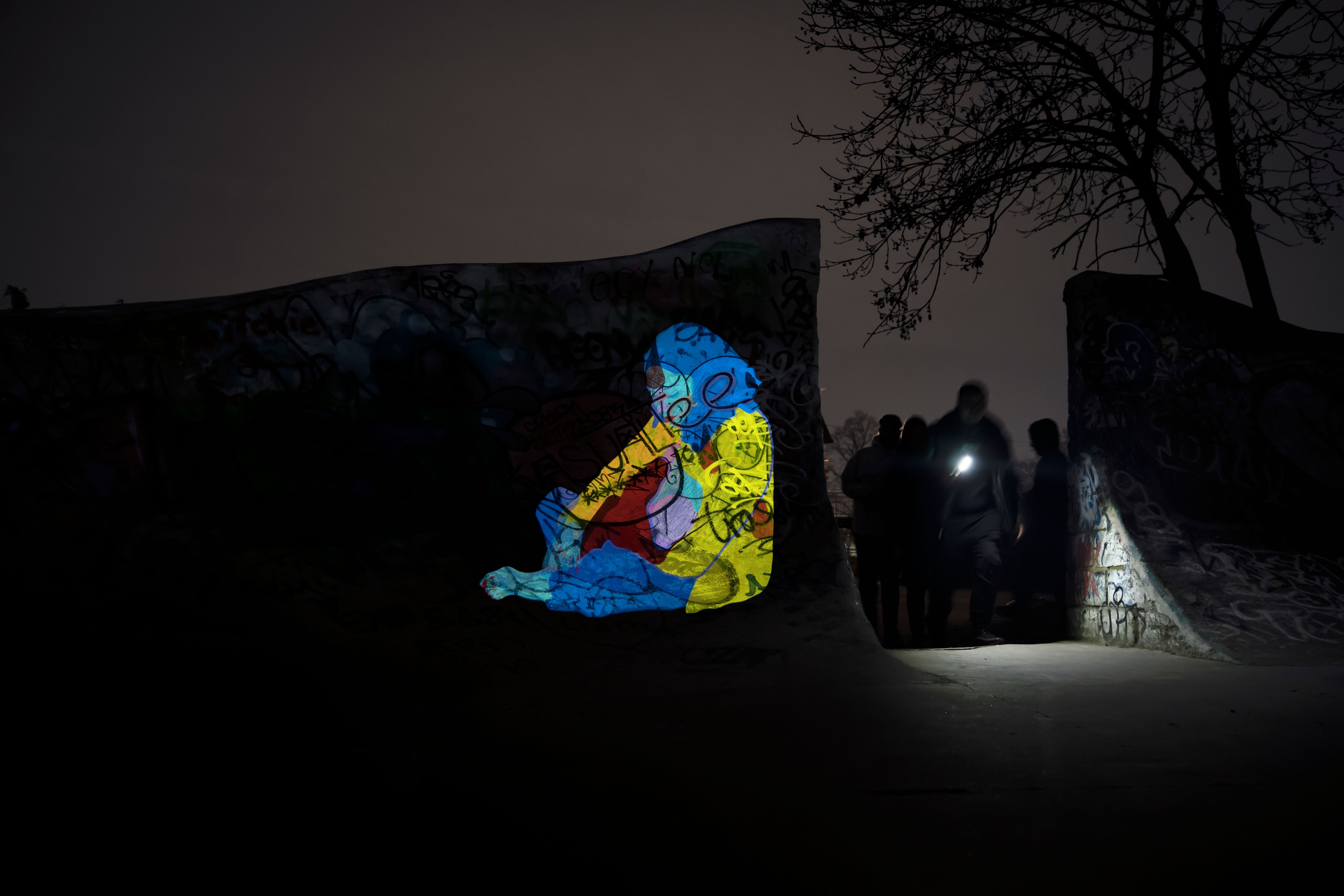 A light projection shows a seated figure as a group of people walk past with a torch.