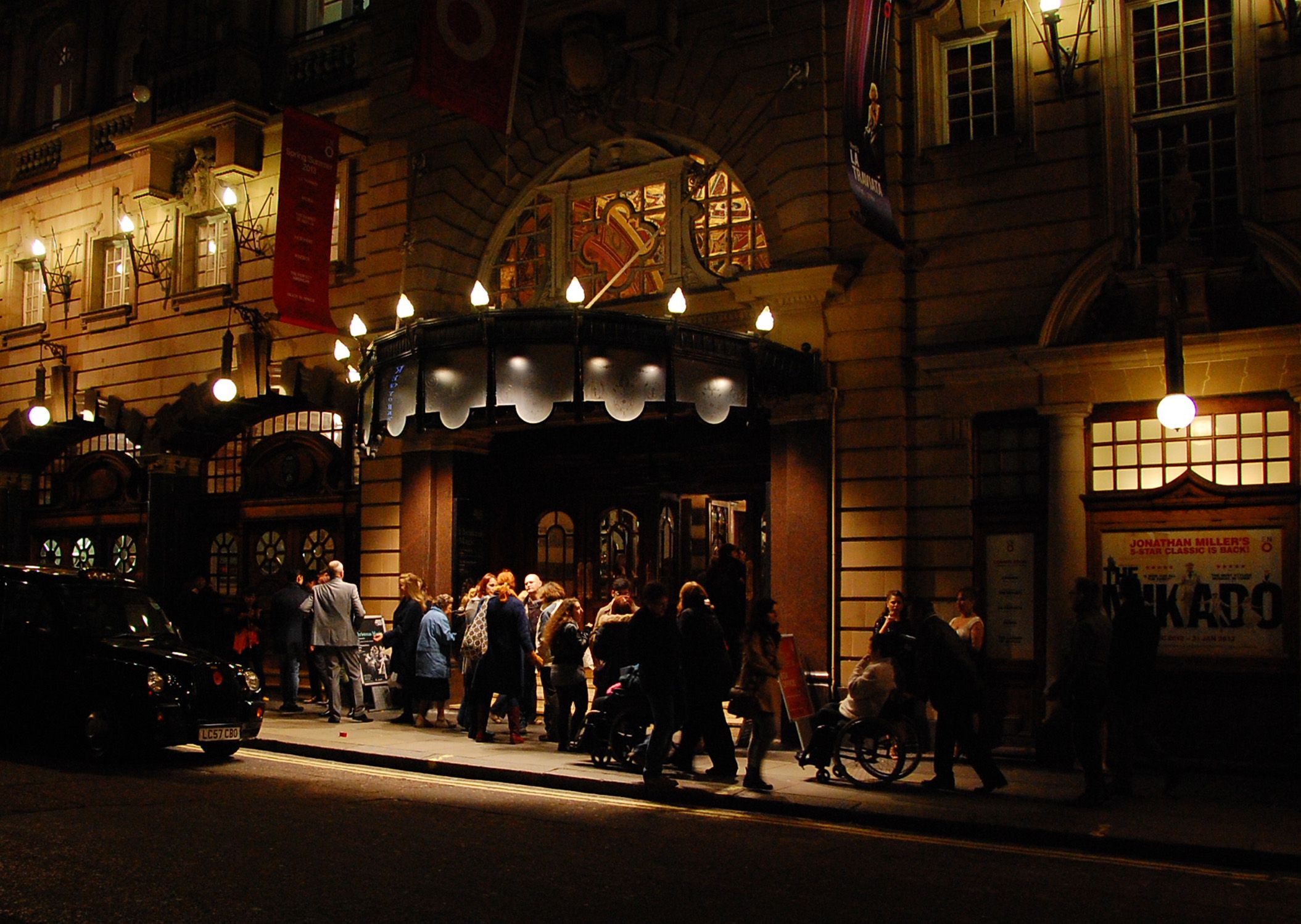 People waiting beneath an illuminated theatre portico.