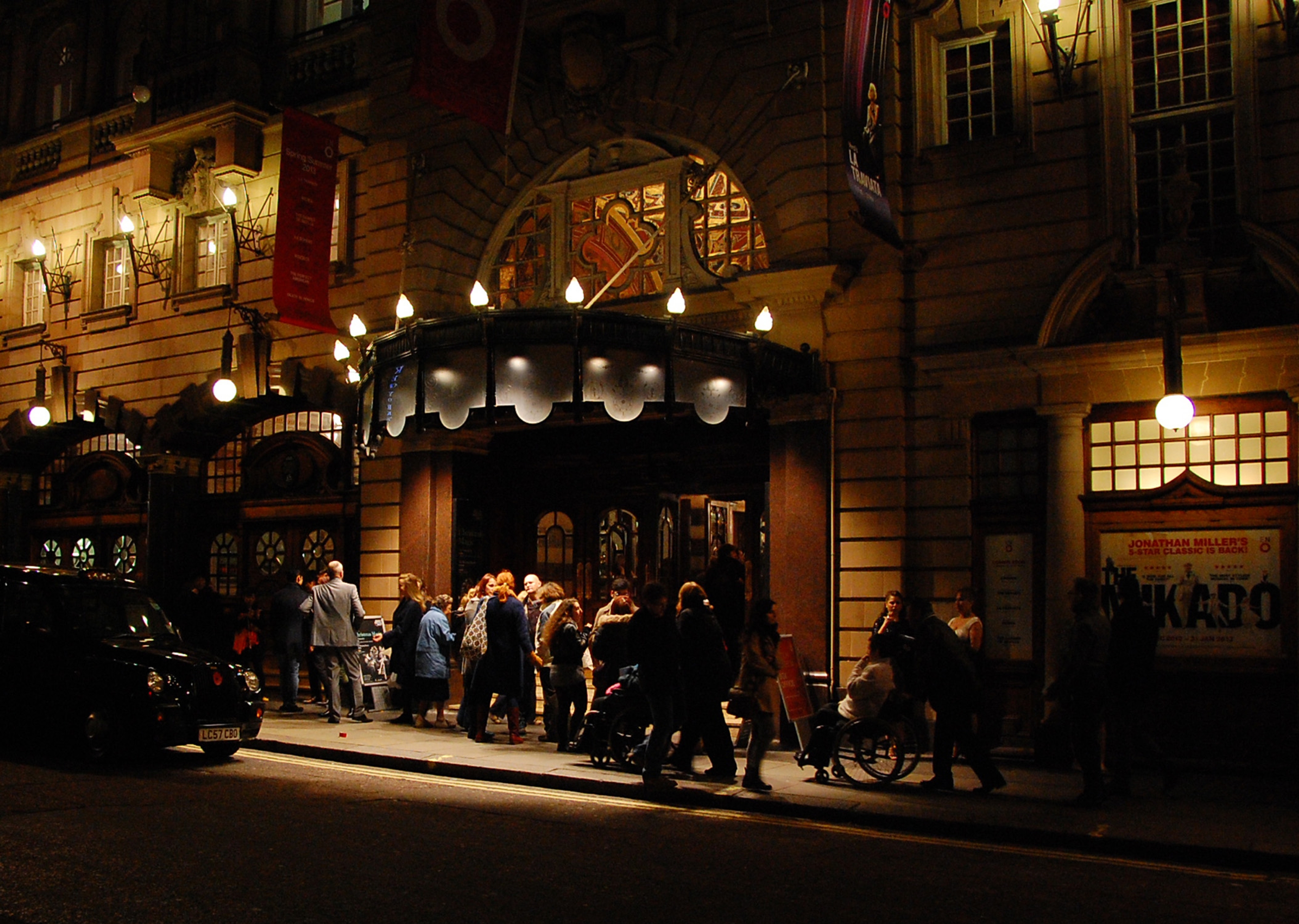People waiting beneath an illuminated theatre portico.