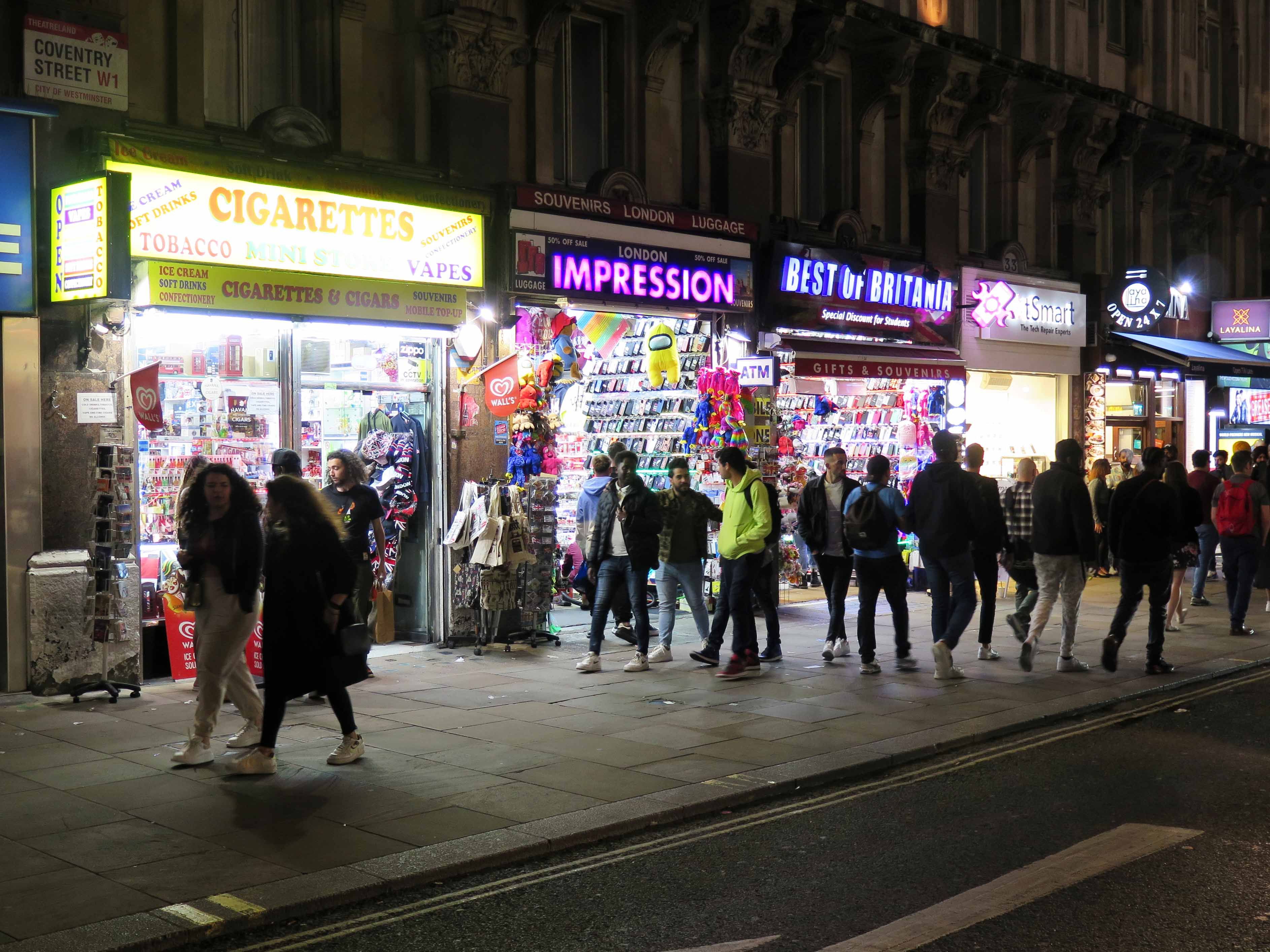 Pedestrians walking past brightly lit shop interiors and signs at night.