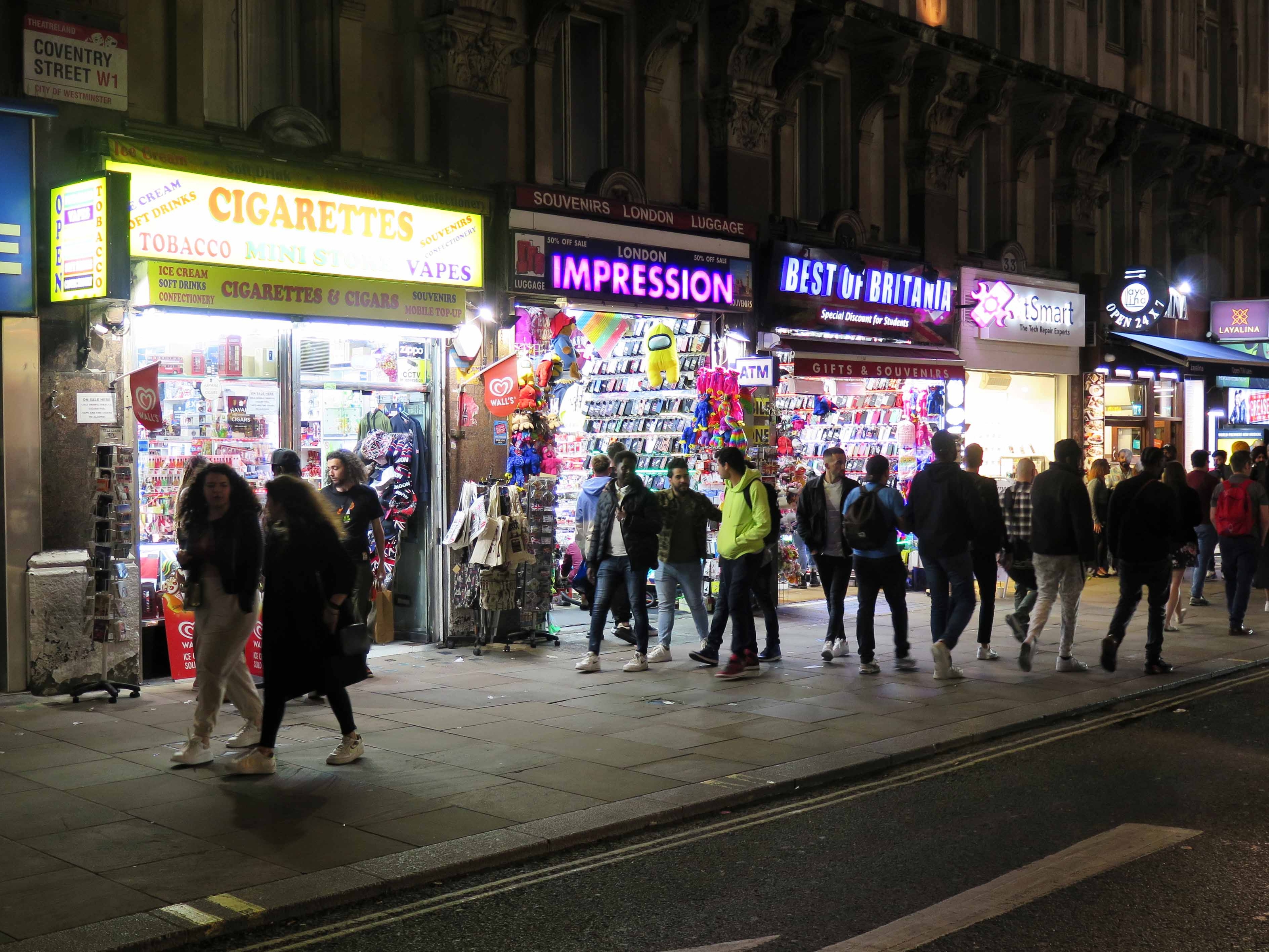 Pedestrians walking past brightly lit shop interiors and signs at night.