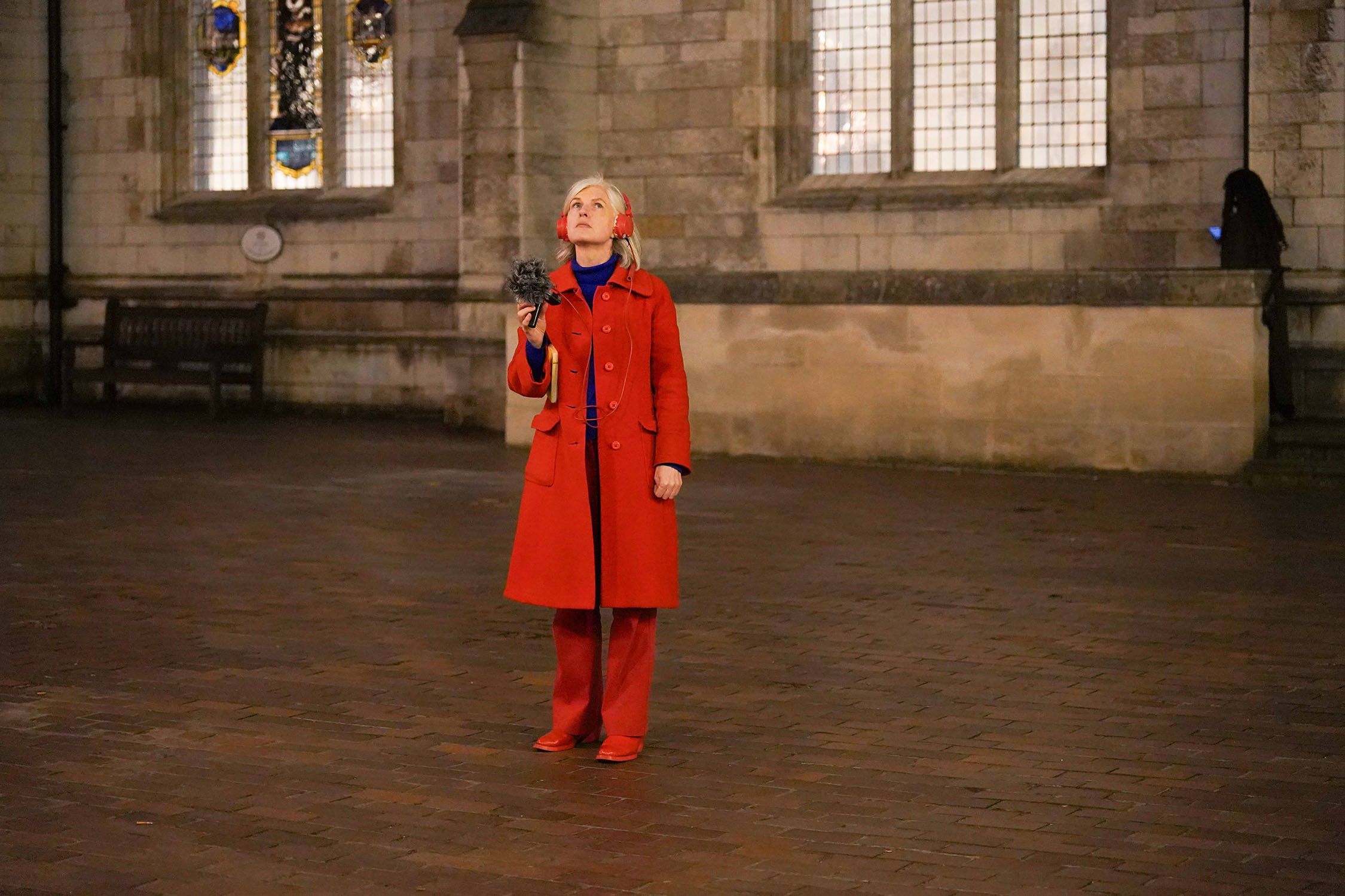 A person wearing a bright red jacket and boots looks up while holding up a microphone and wearing red headphones.