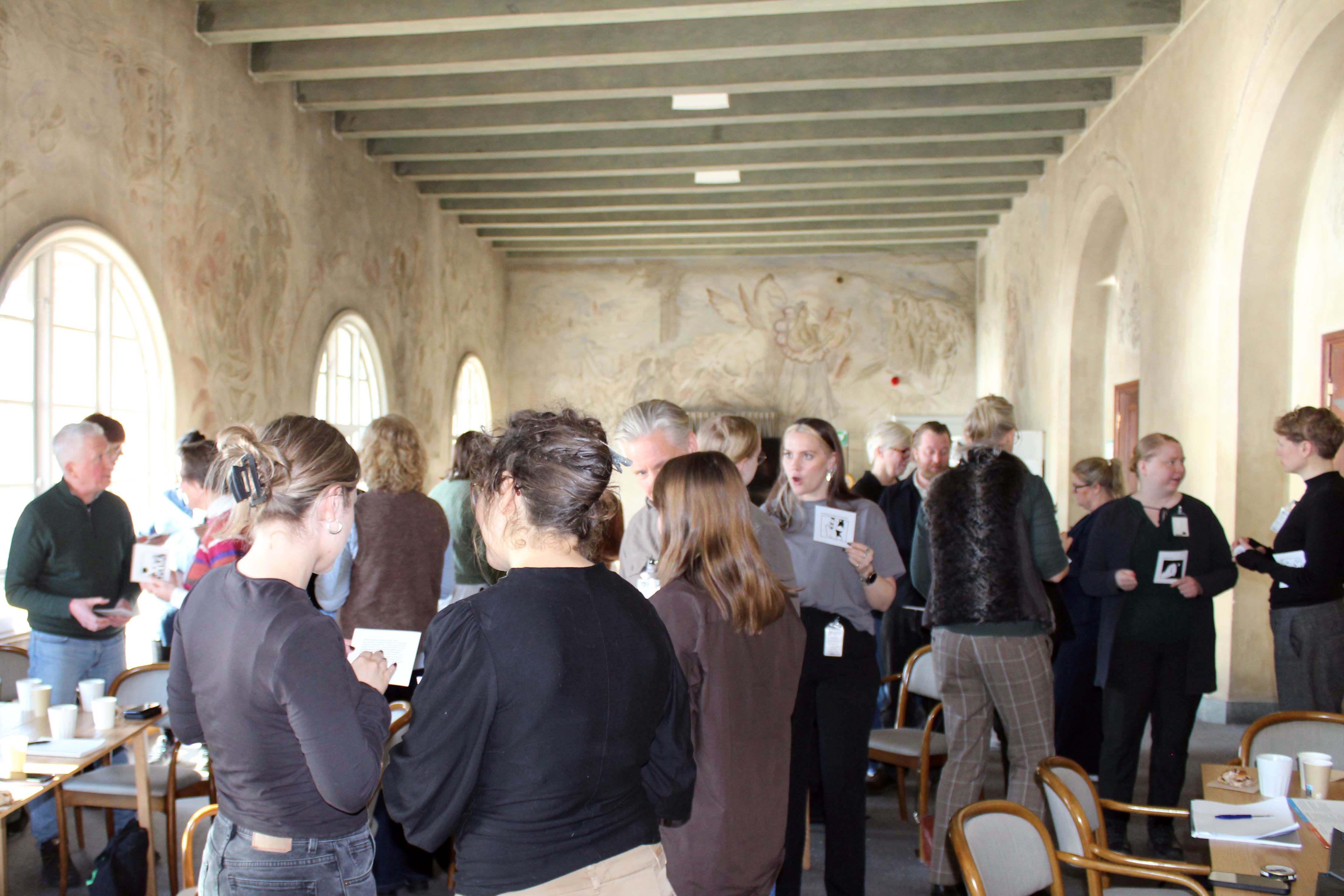 A large group stands in a room talkin gin small groups and holding up small printed cards.