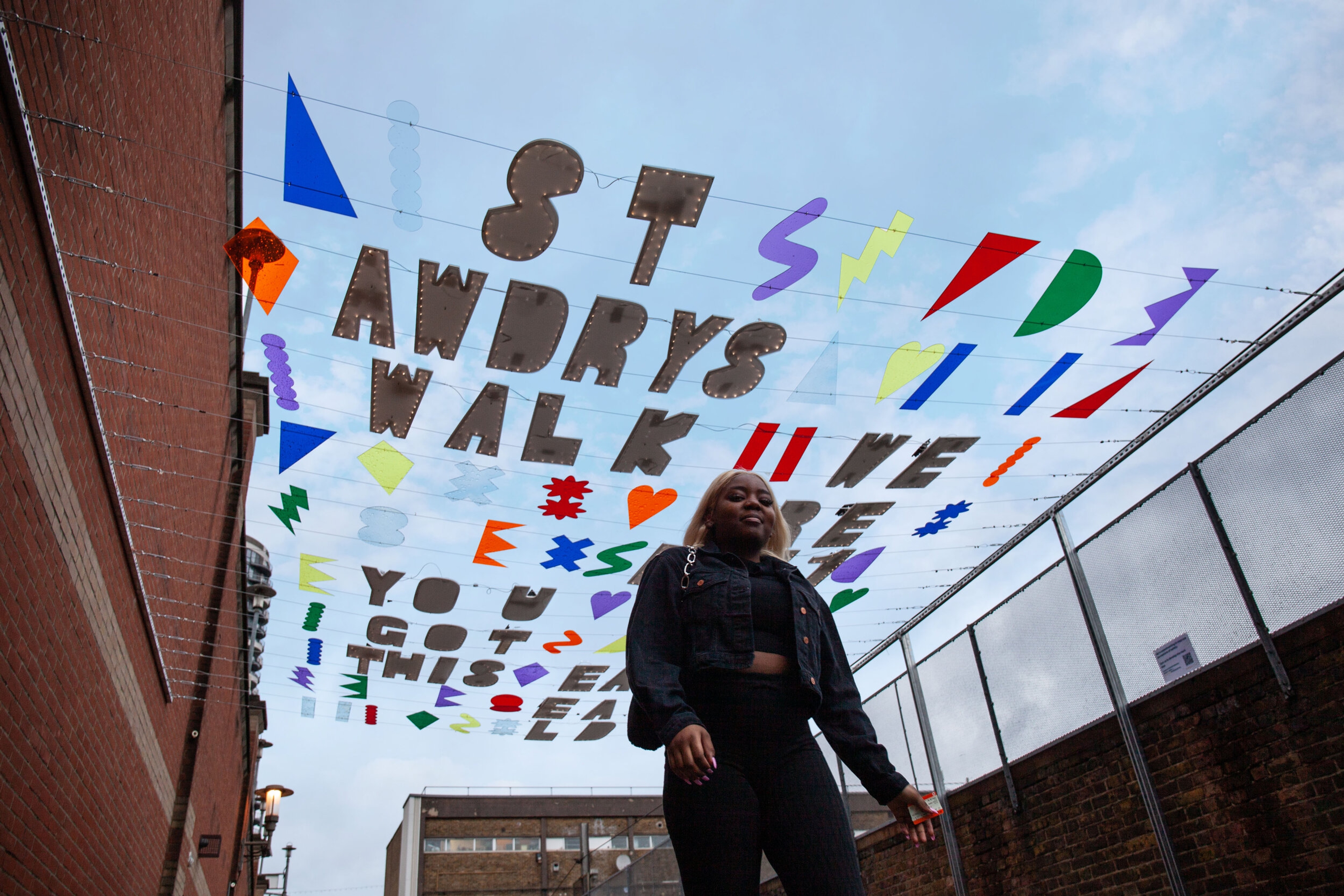 A person walks beneath colourful text and shapes suspended between two buildings above a street.
