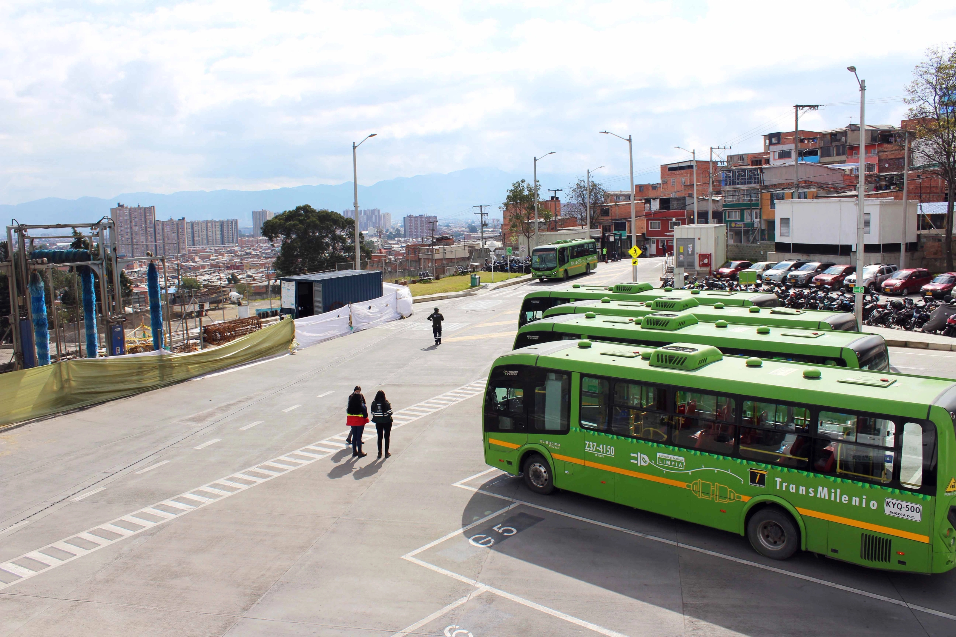 Four green buses are parked in a large outdoor bus depot.