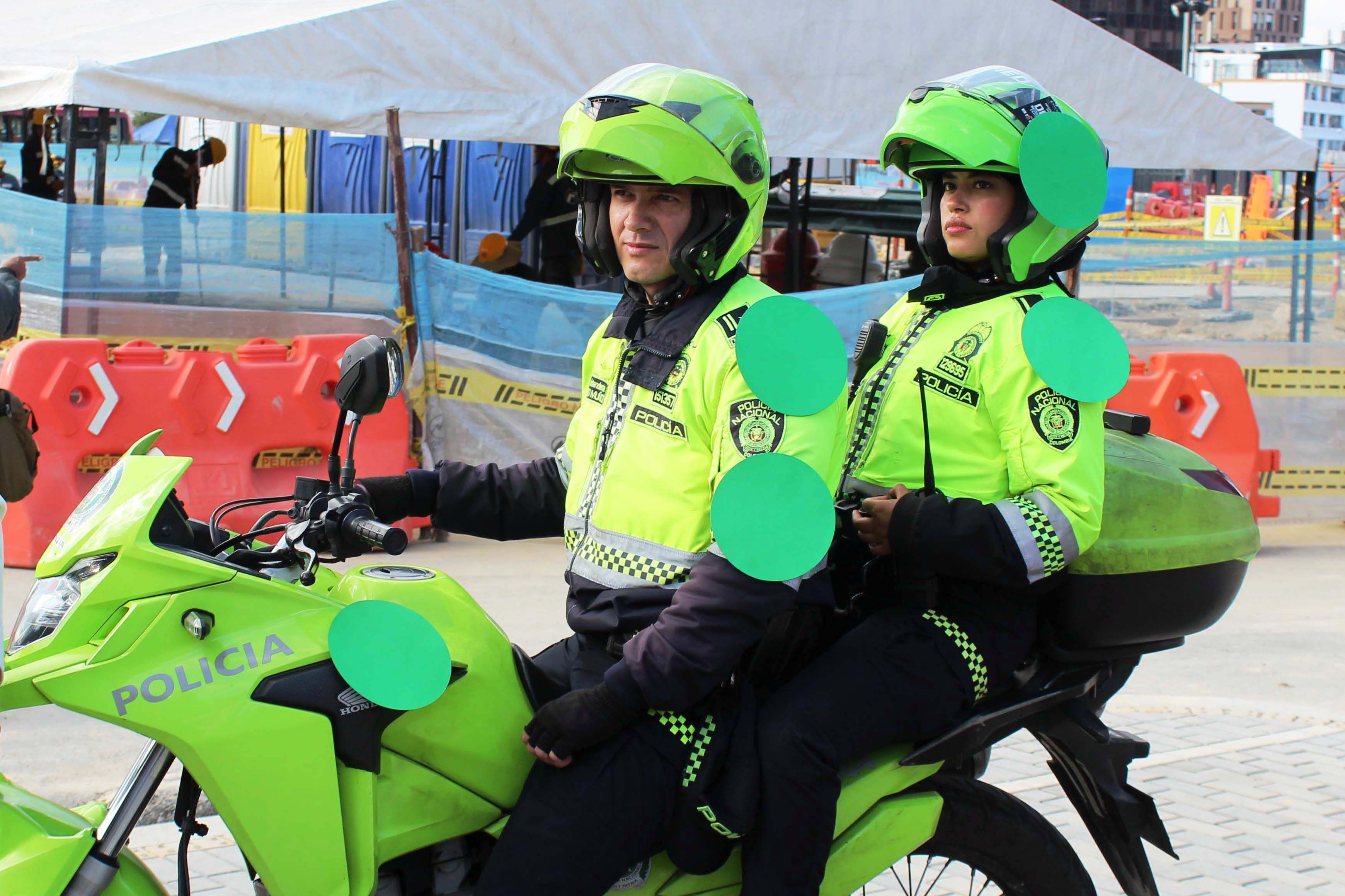 Two police officers sit on a motorcycle wearing hi-vis jackets and helmets. The police officers are have four green paper circles stuck to them.