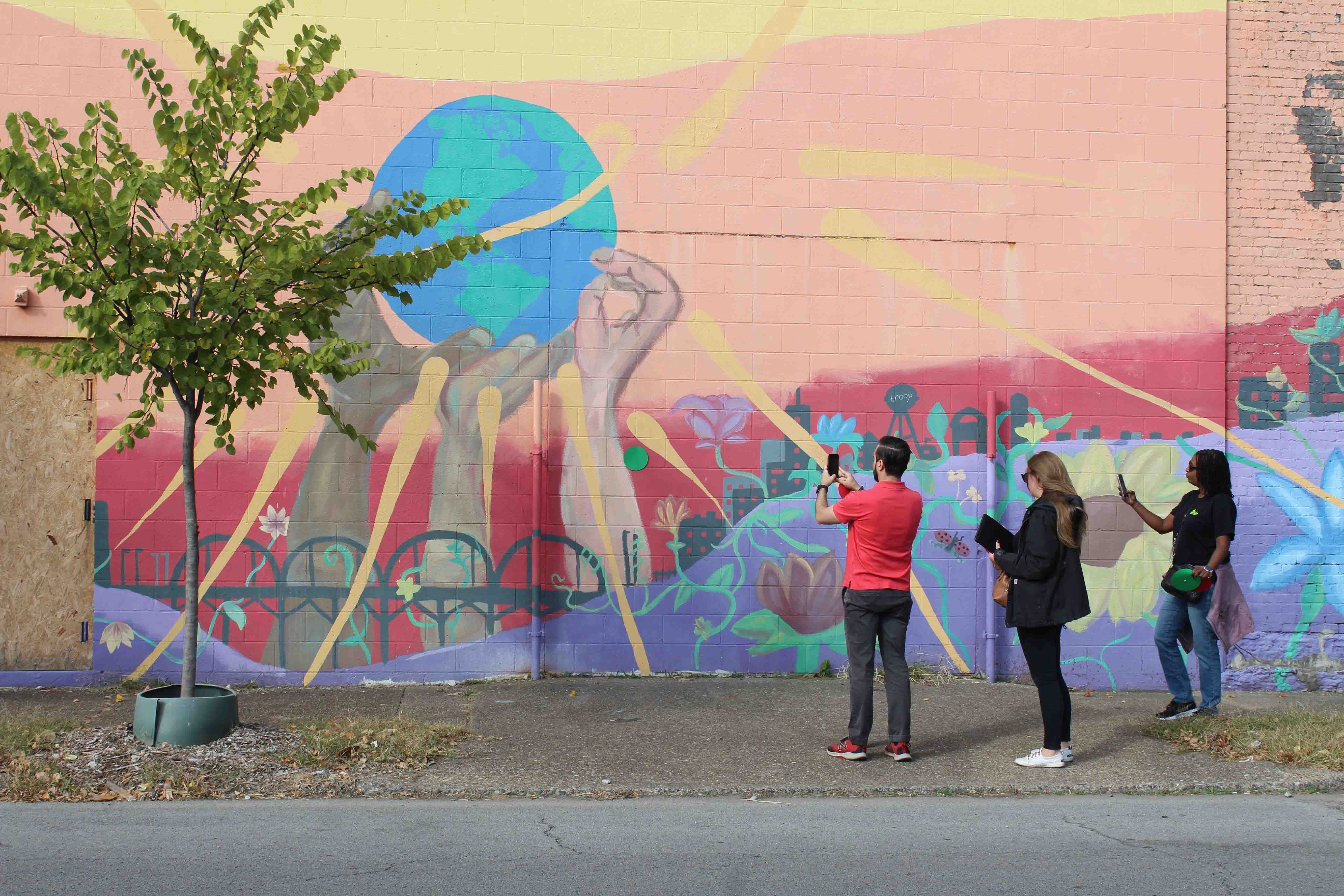 Three people take a photograph of a colourful mural showing three hands holding up the earth.