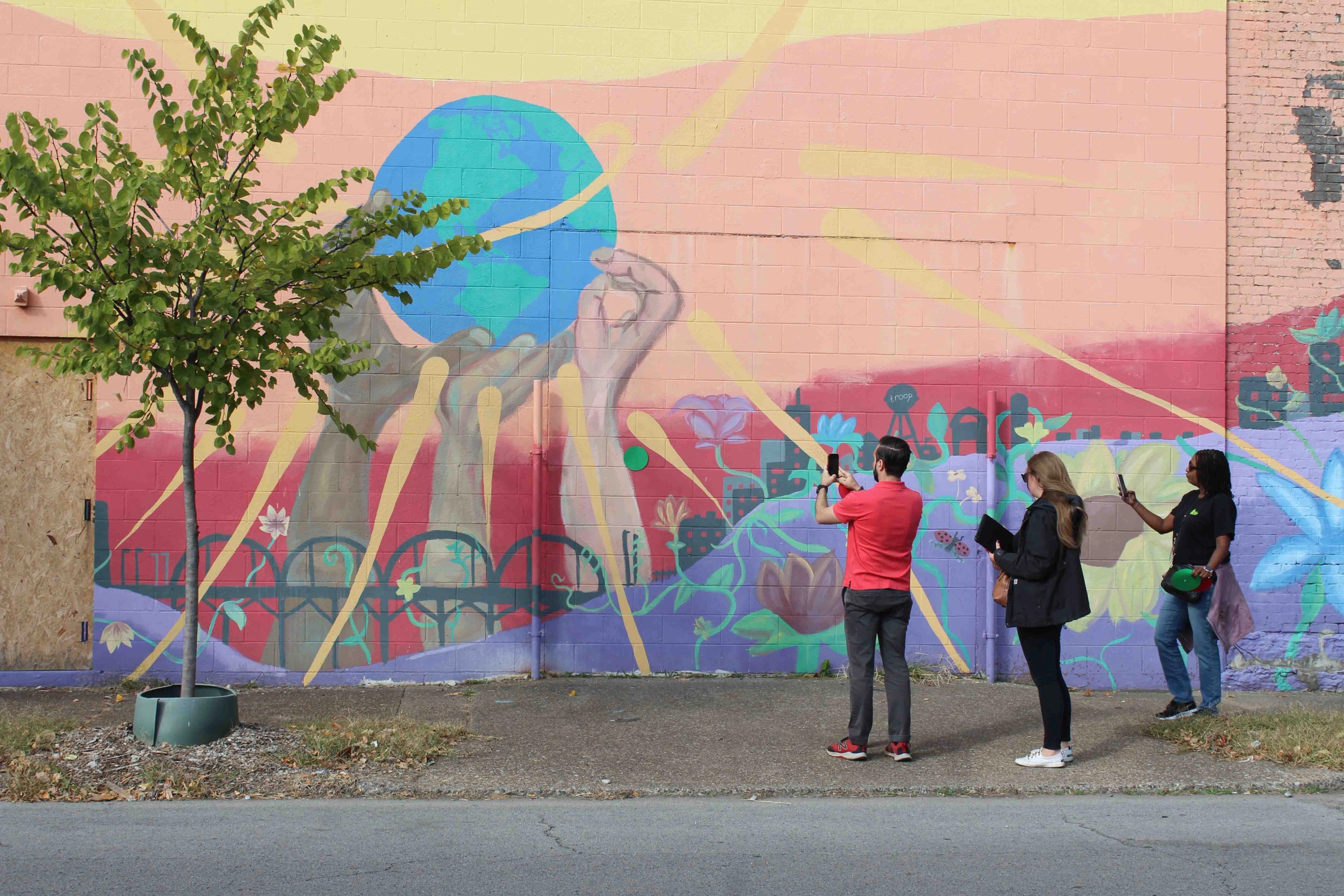 Three people take a photograph of a colourful mural showing three hands holding up the earth.