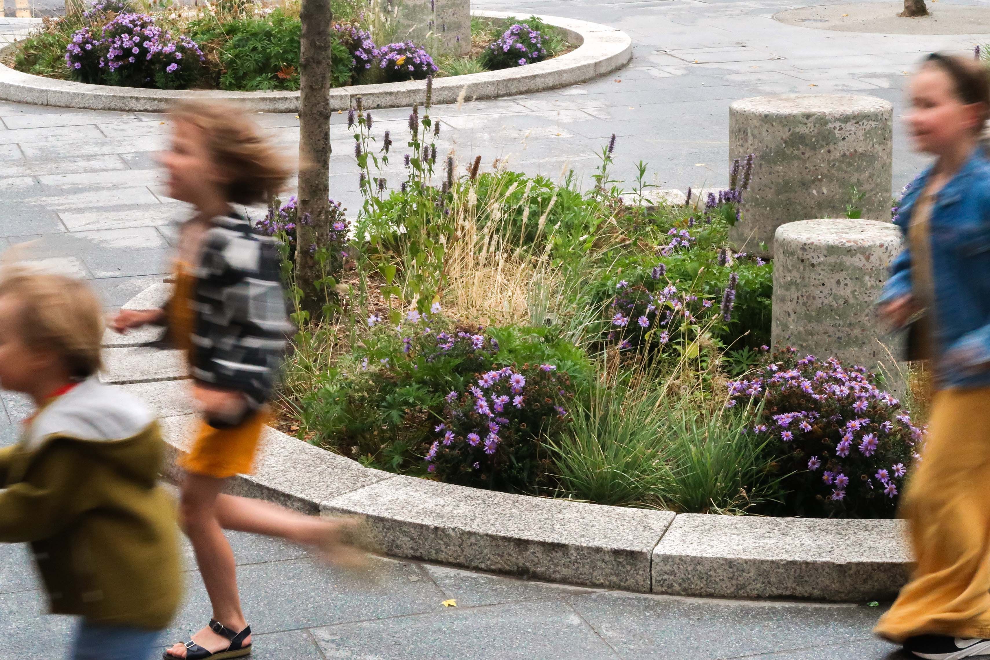 Children running past a flower bed on a pavement.