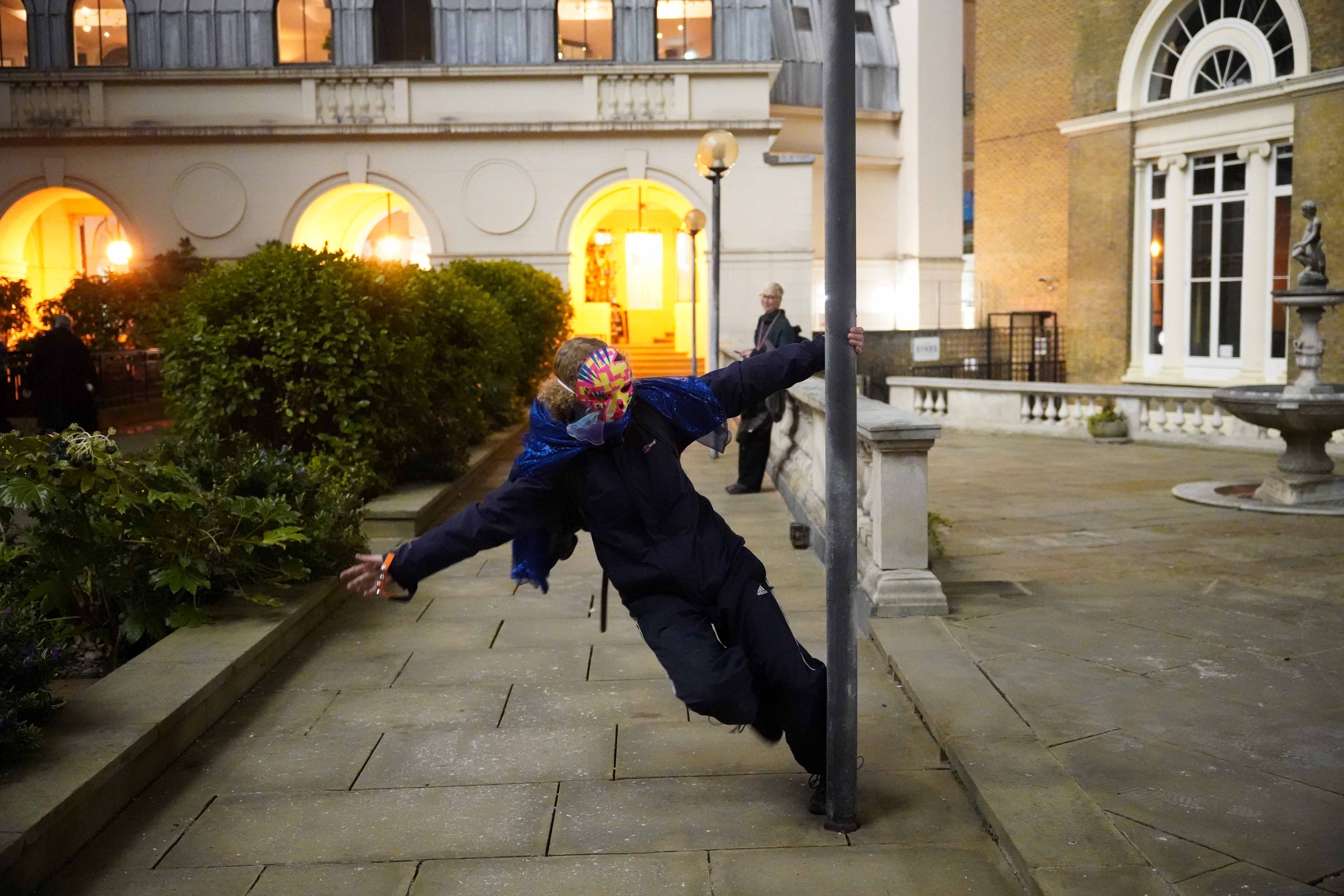A person wearing a decorated mask holds onto a lamp column with one hand and leans over away from the column in a courtyard.