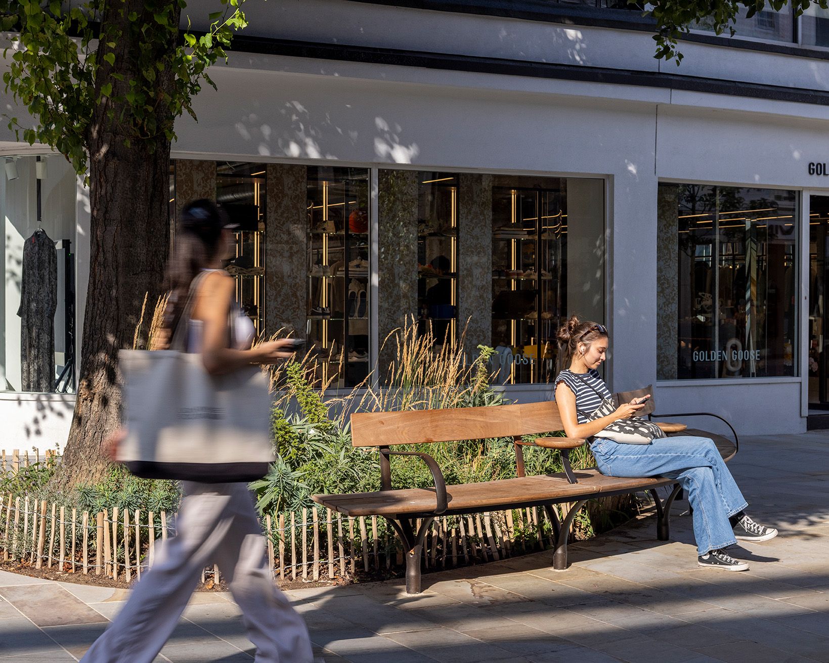 A person sits on a bench with their back to a tree and planted area bordered by a small wooden fence. 
