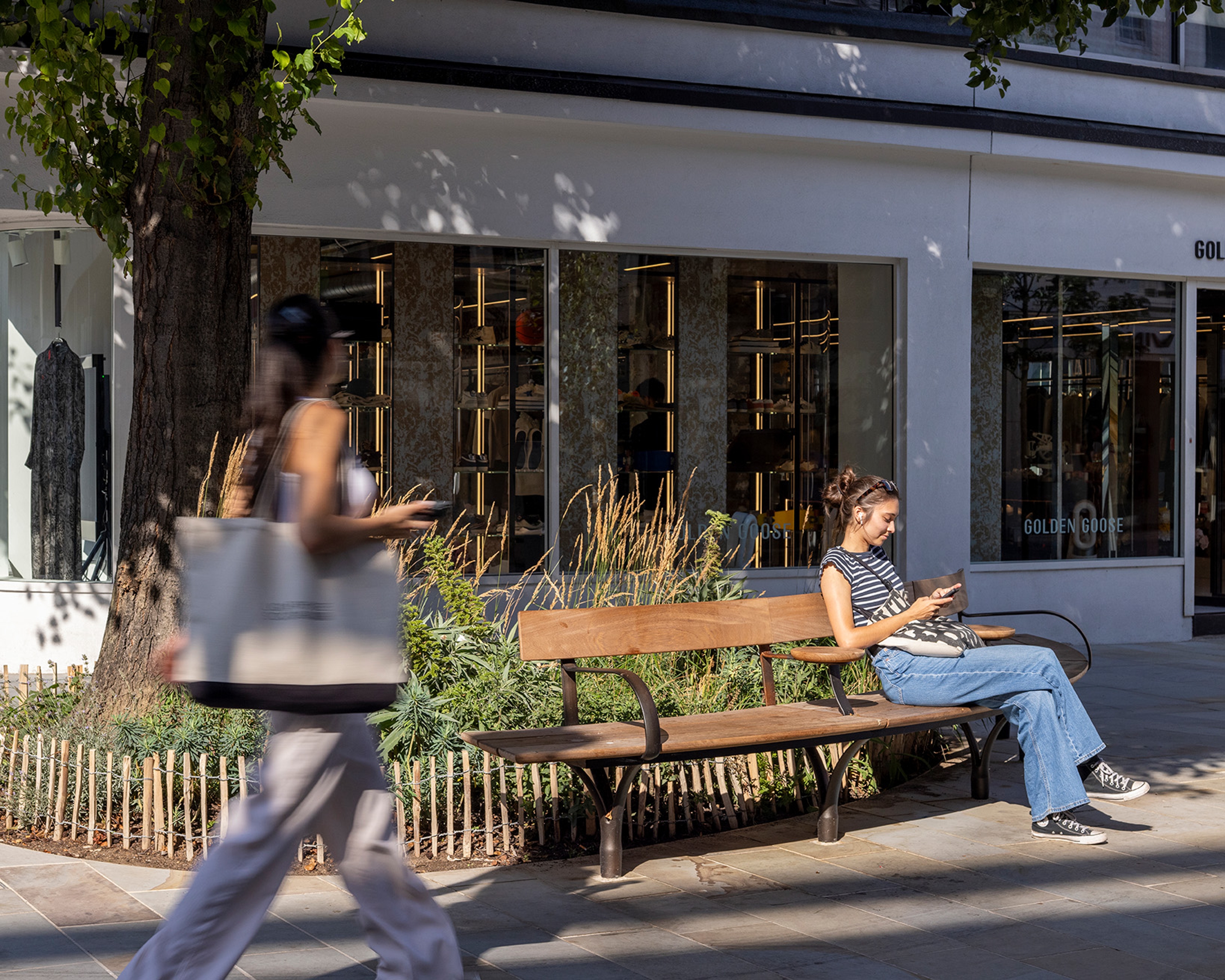 A person sits on a bench with their back to a tree and planted area bordered by a small wooden fence.