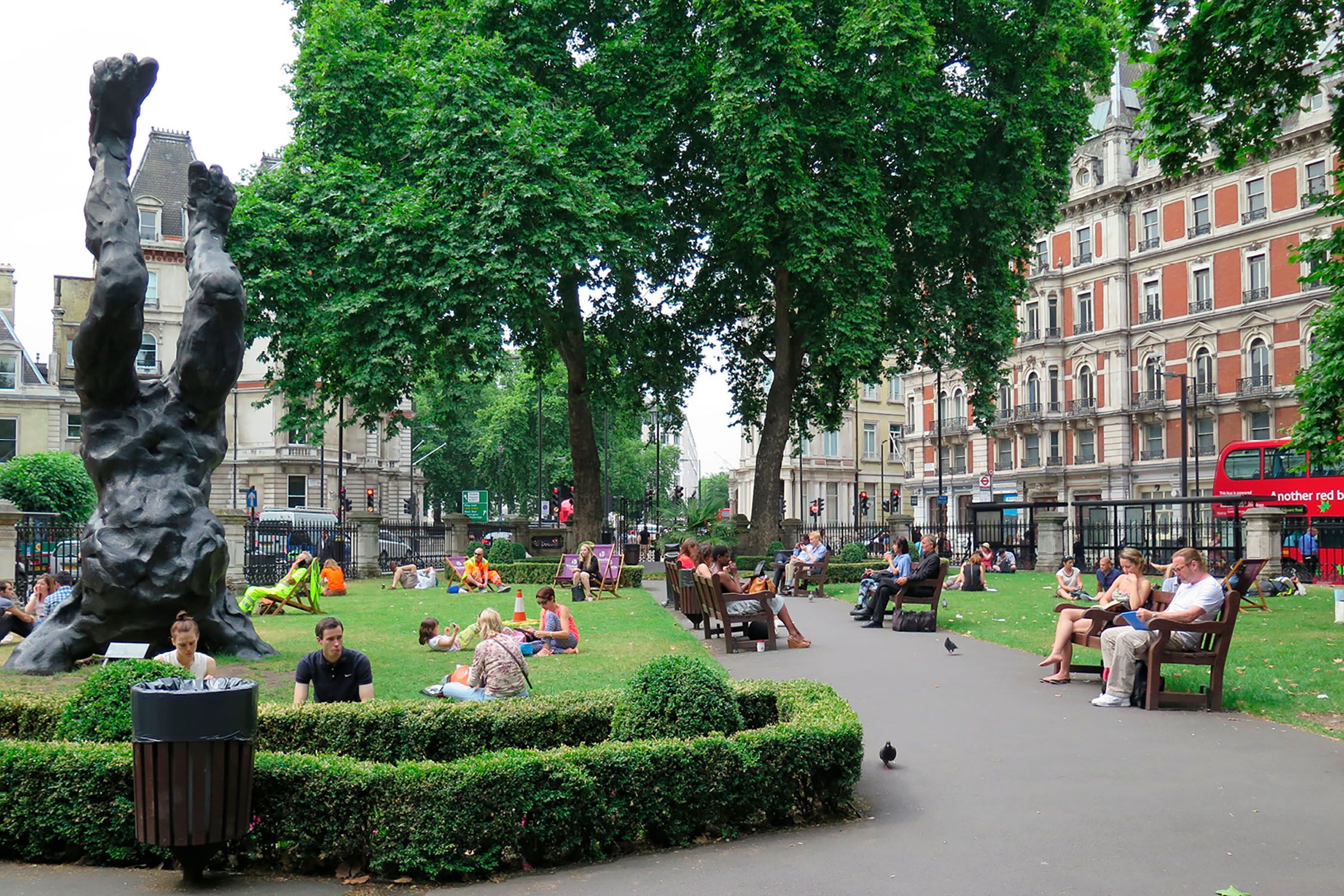 A garden square with a large sculpture and people sitting on the grass and on benches.