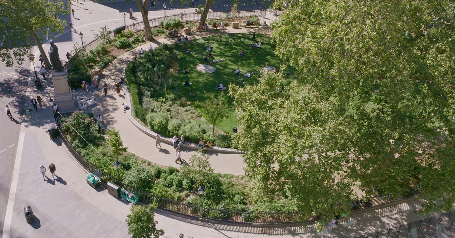 Photograph of the edge of Hanover Square taken from above with green bushes and trees and people walking on the pavement around it.