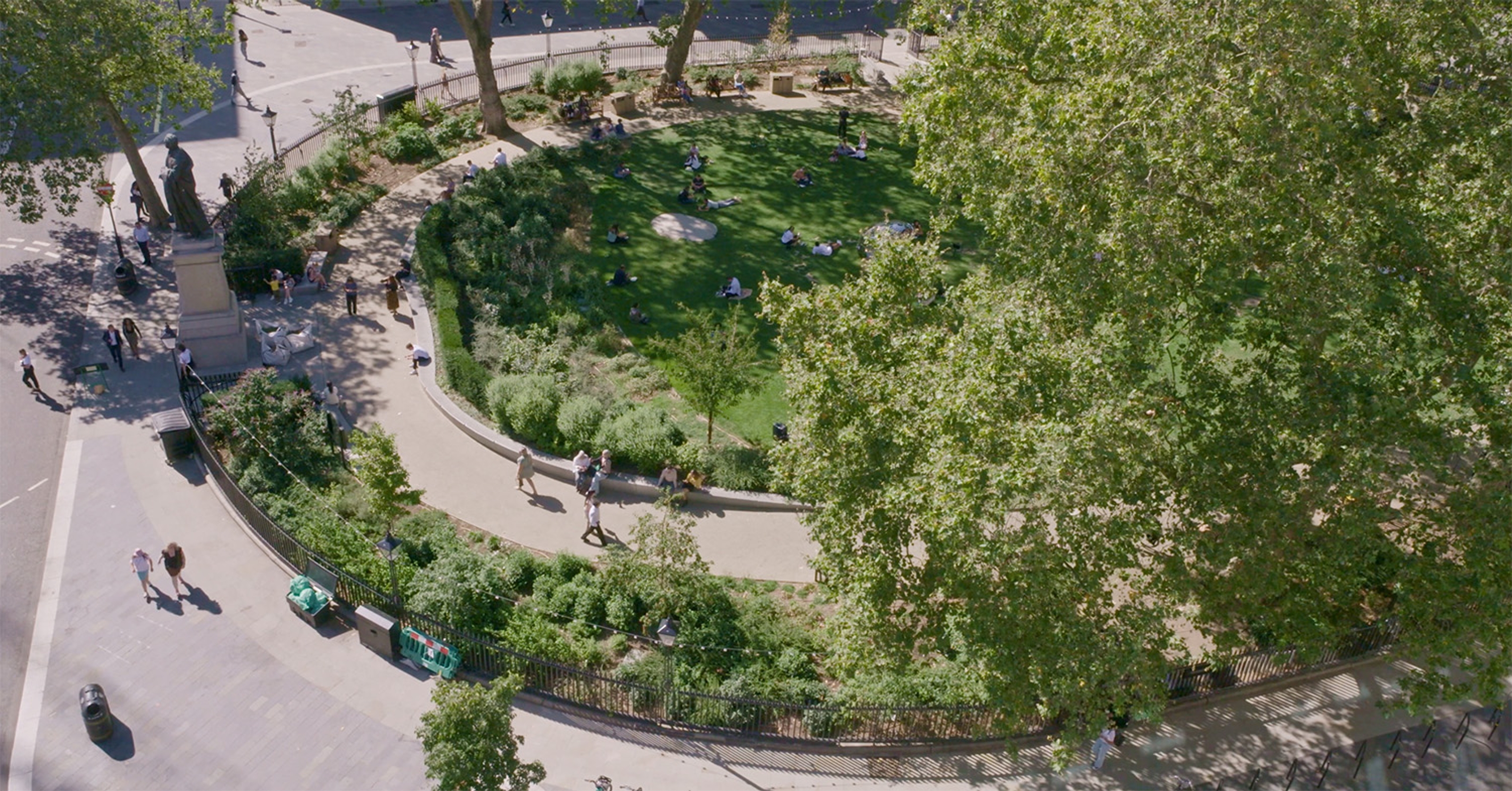 Photograph of the edge of Hanover Square taken from above with green bushes and trees and people walking on the pavement around it.