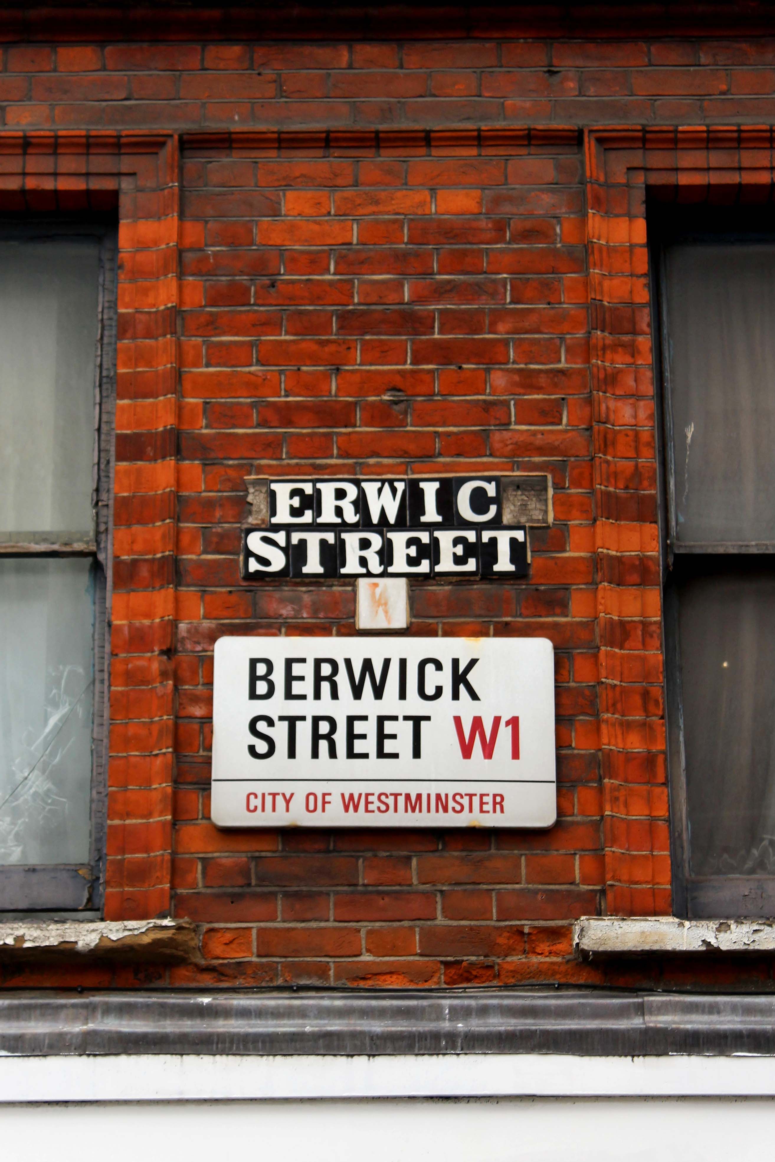 A modern street sign underneath historic letter tiles of the street name with the letters b and k missing from Berwick Street.