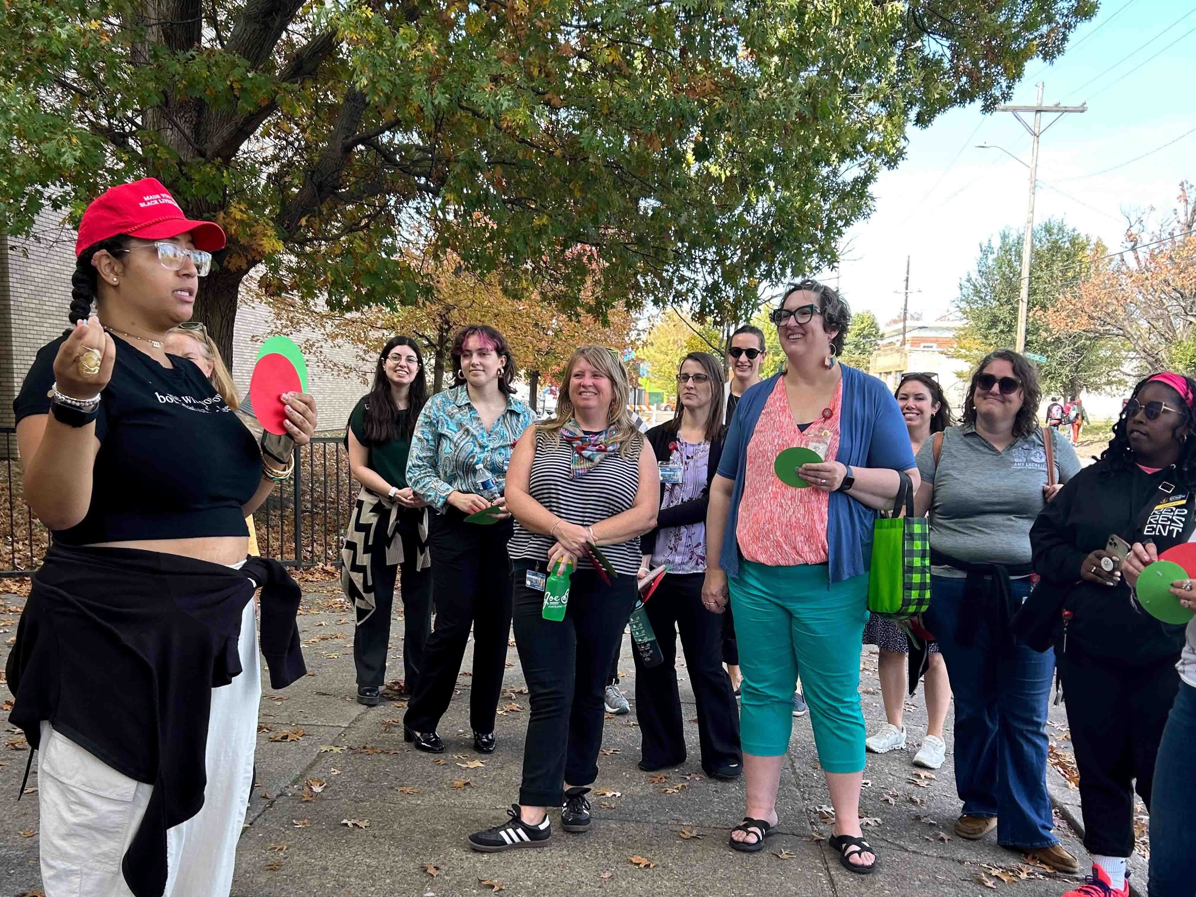 A community activist in a red hat speaks to a group of people on a pavement.
