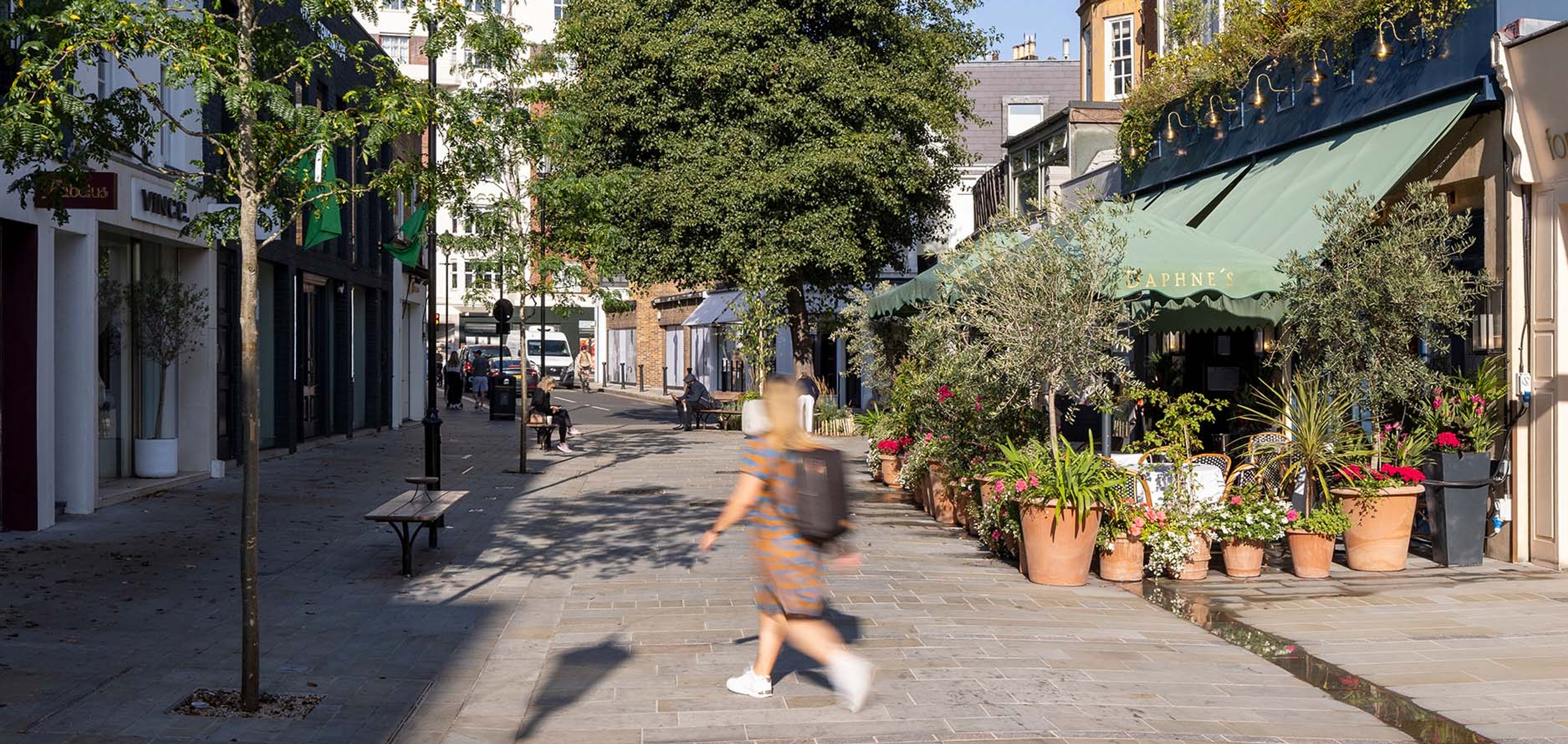 A person is walking across Draycott Avenue with a tree in the centre and shops lining the street.