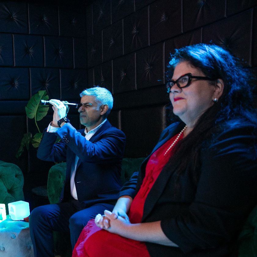 Amy Lamé and Sadiq Khan at a nighttime event indoors, watching something out of shot.