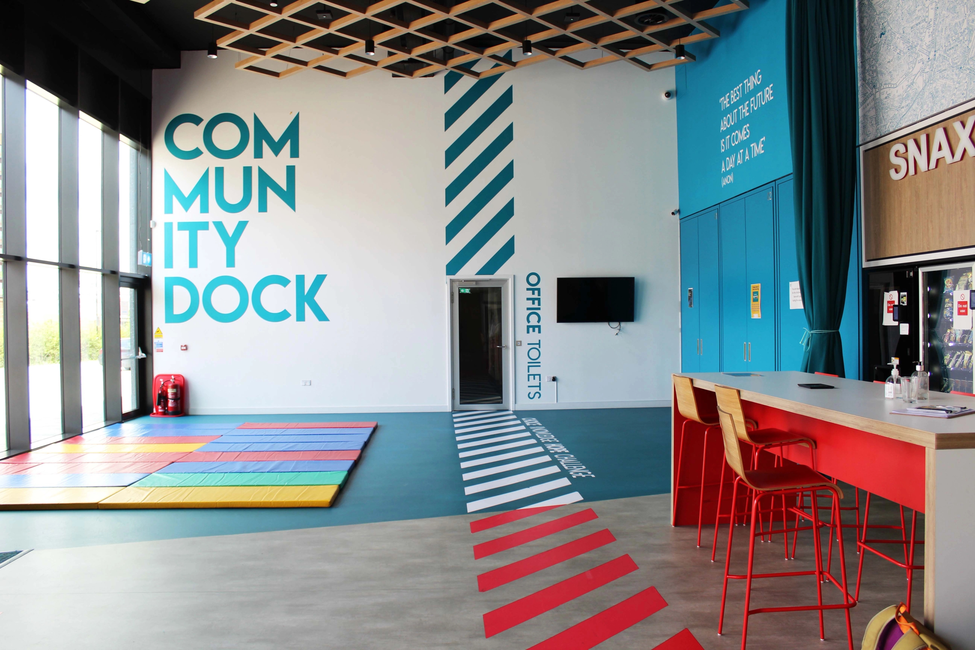 A community space with high ceilings, soft mats on the floor, tables and chairs and large double height windows.