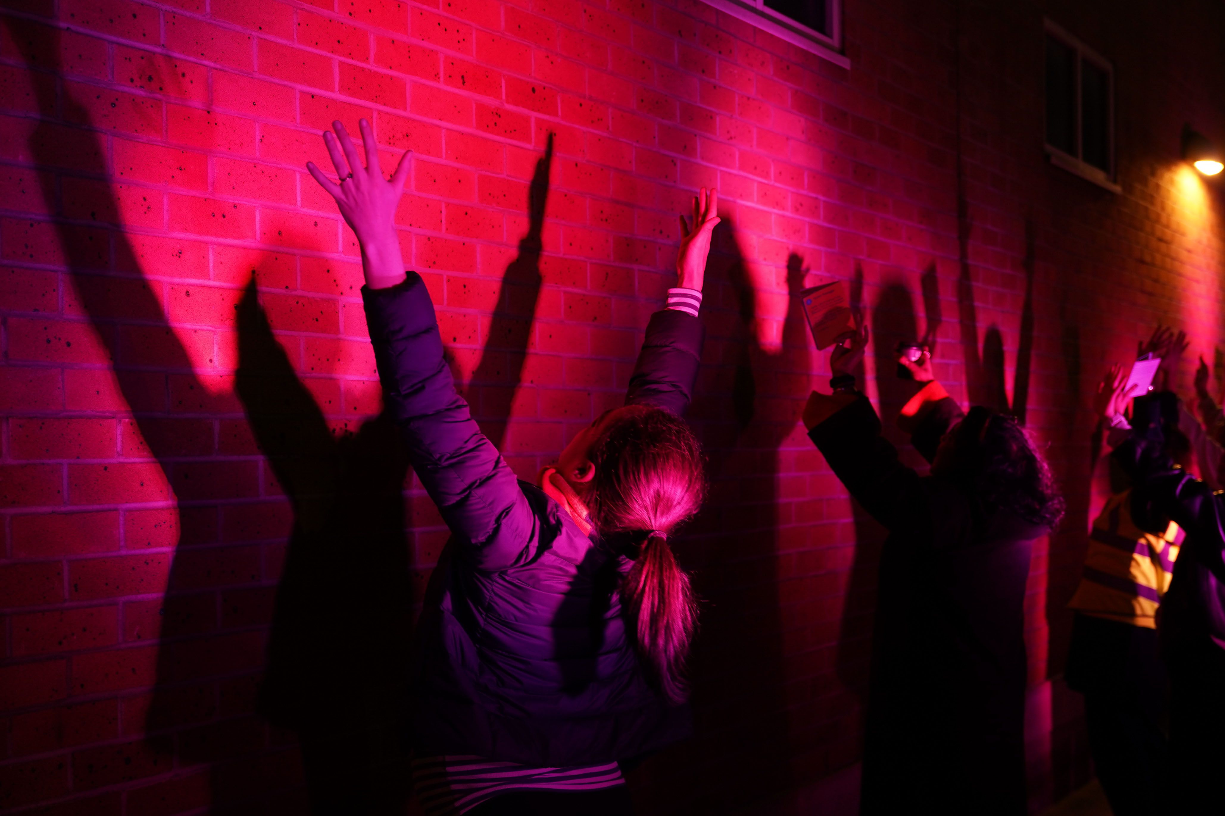 Two people hold their arms up while facing a wall as pink light is projected onto the walls, creating shadows. 