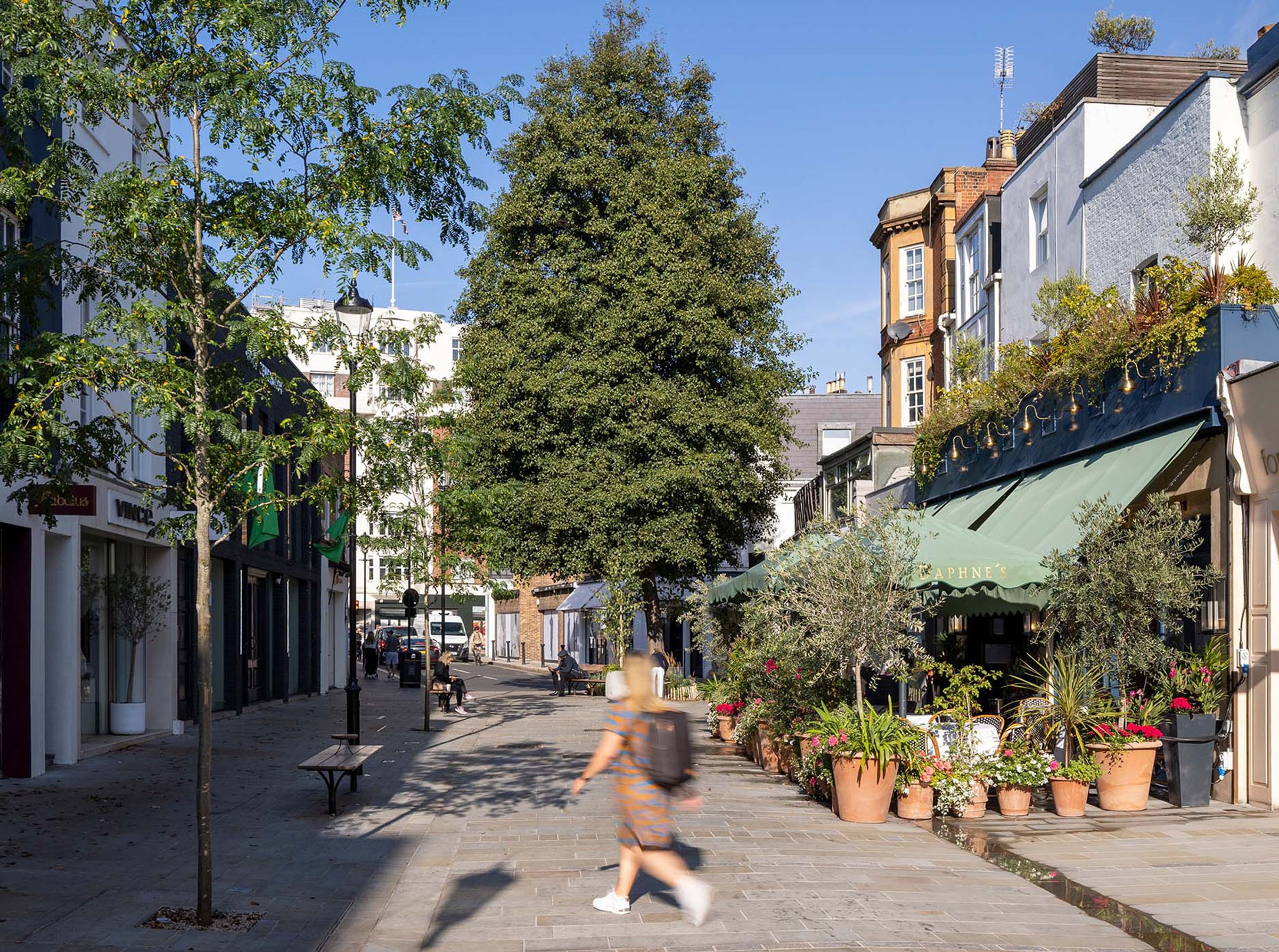 A person is walking across Draycott Avenue with a tree in the centre and shops lining the street.