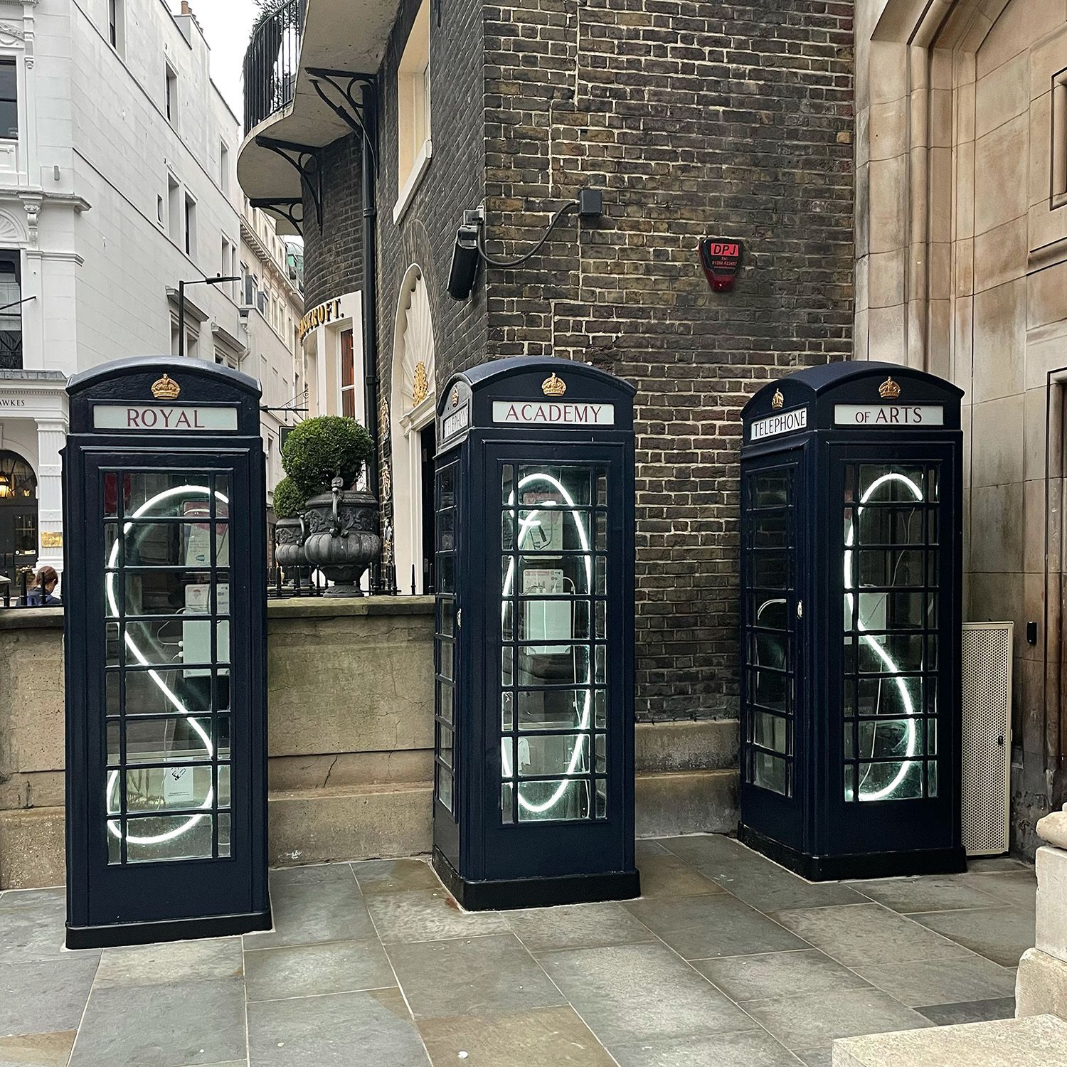 Three black telephone box art installations with glowing white neon letters reading "SOS" displayed outside the Royal Academy of Arts on Burlington Gardens.
