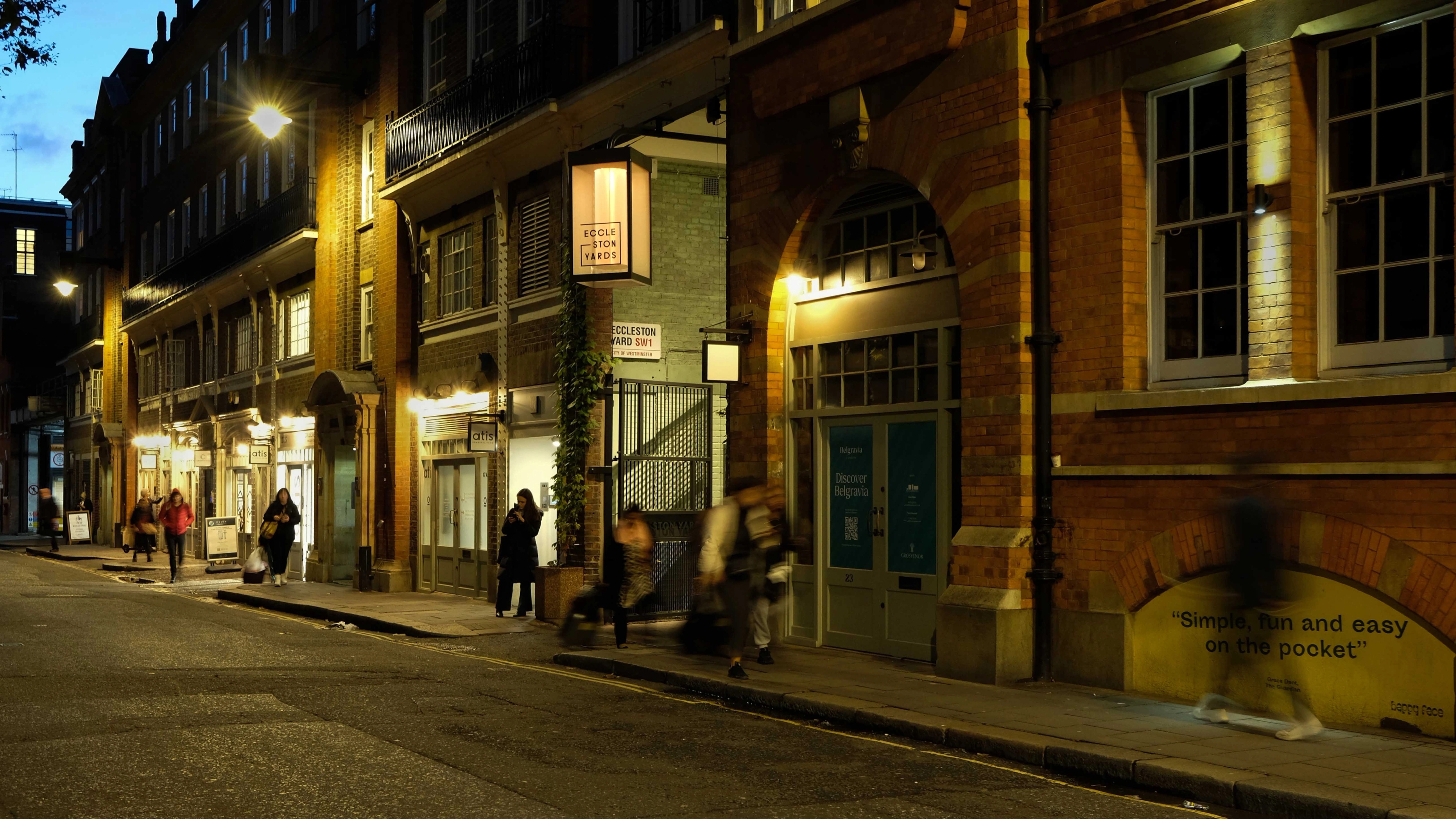 The entrance to Eccleston Yards after dark with a large illuminated lamp announcing the entrance to the yard as people walk past on the pavement.