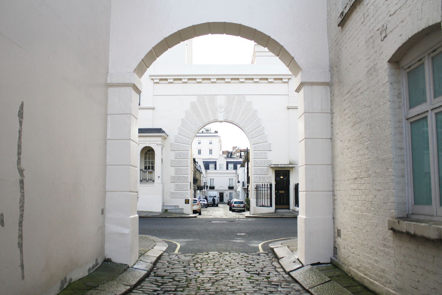 A view through two archways looking towards a cobbled Mews street with cars parked.