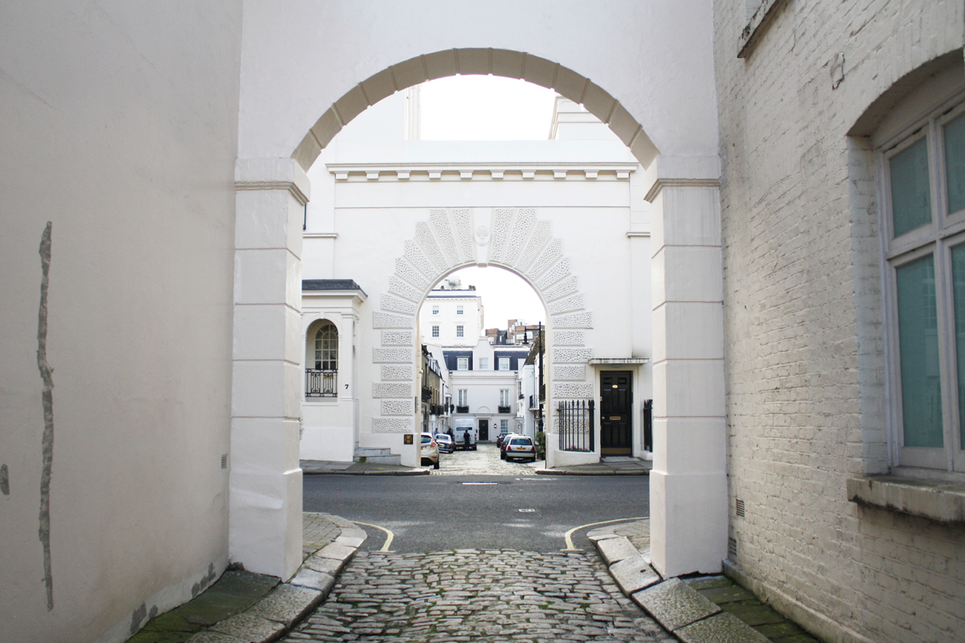 A view through two archways looking towards a cobbled Mews street with cars parked.