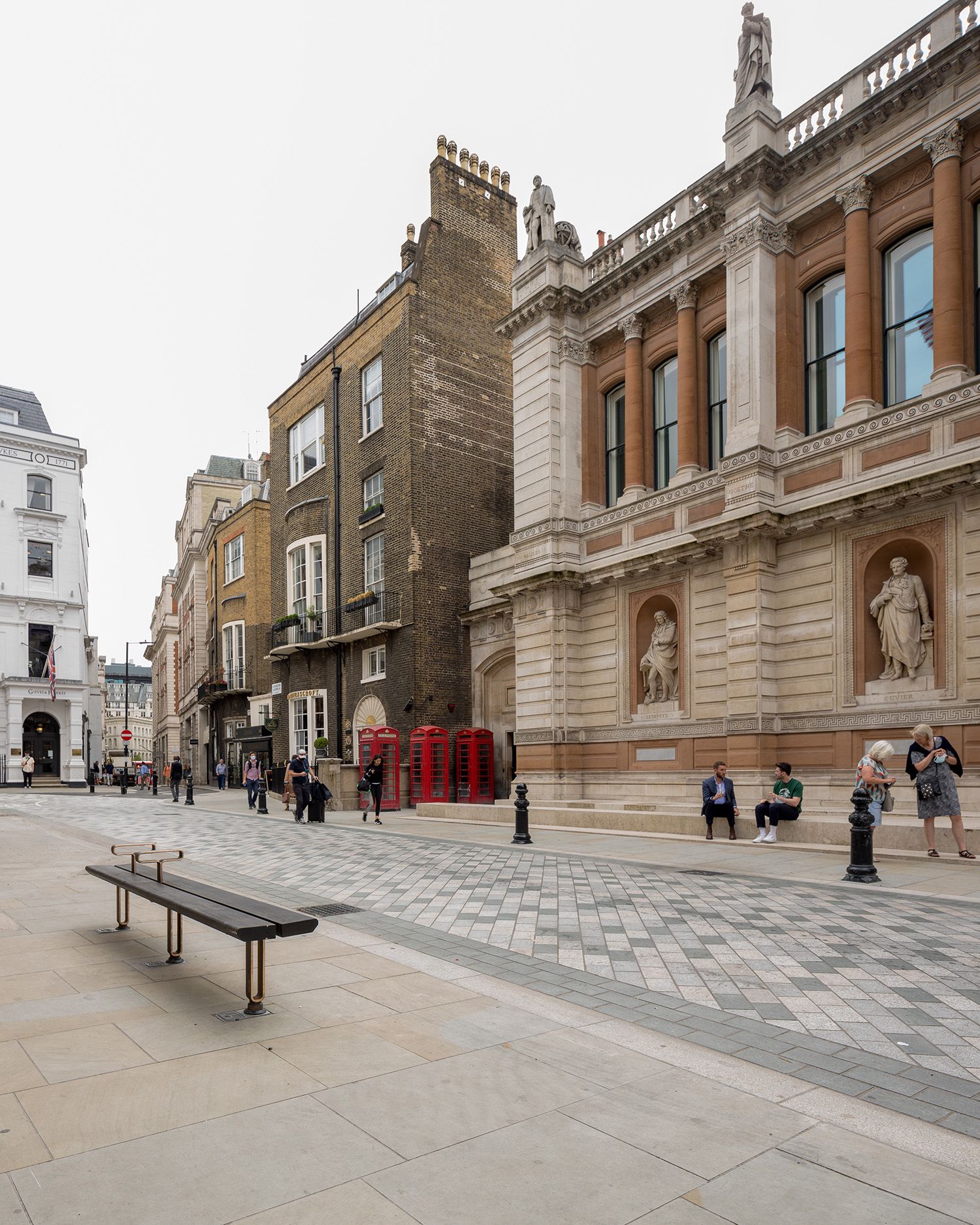 An empty bench on one side of the road with people sitting on the steps of the Royal Academy on the other side of the road.