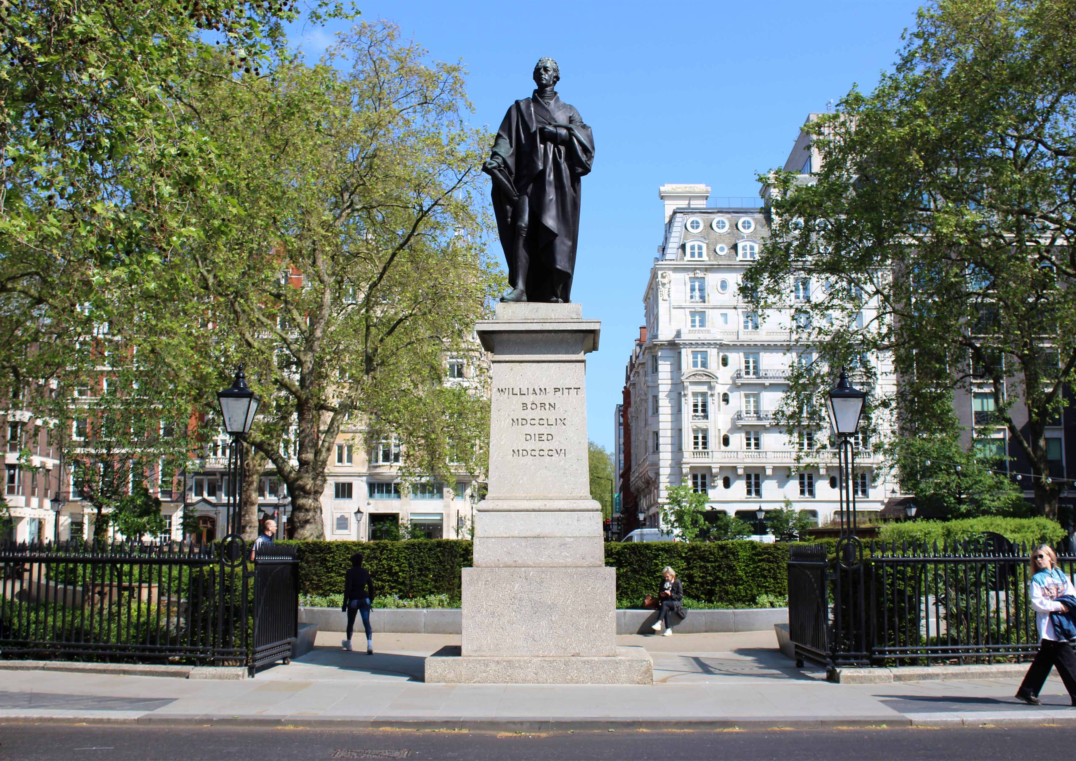 Statue of man on pillar at the entrance to a park with some buildings in the background and a few pedestrians.