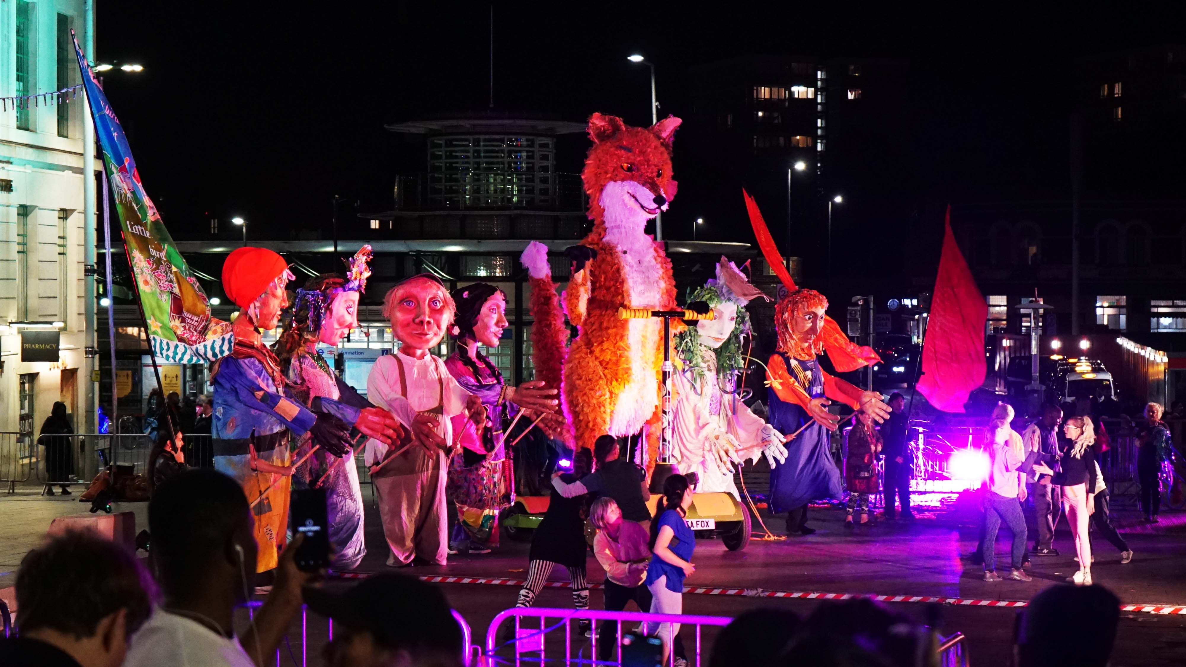 Performers dressed up in colourful paper outfits on a lit up stage at night
