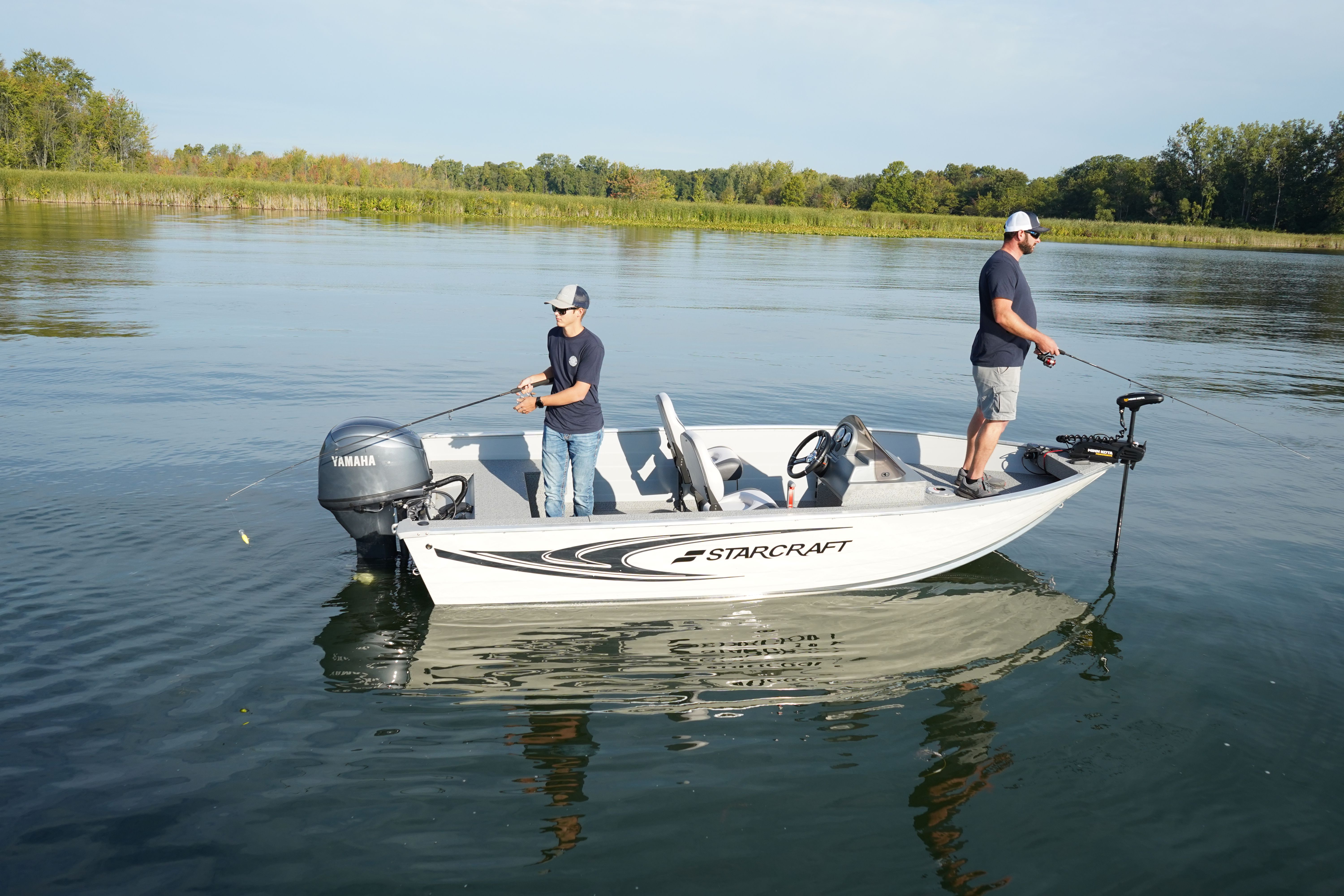 White and grey Starcraft Storm fishing boat with two anglers standing on the deck on calm water near a wooded shoreline