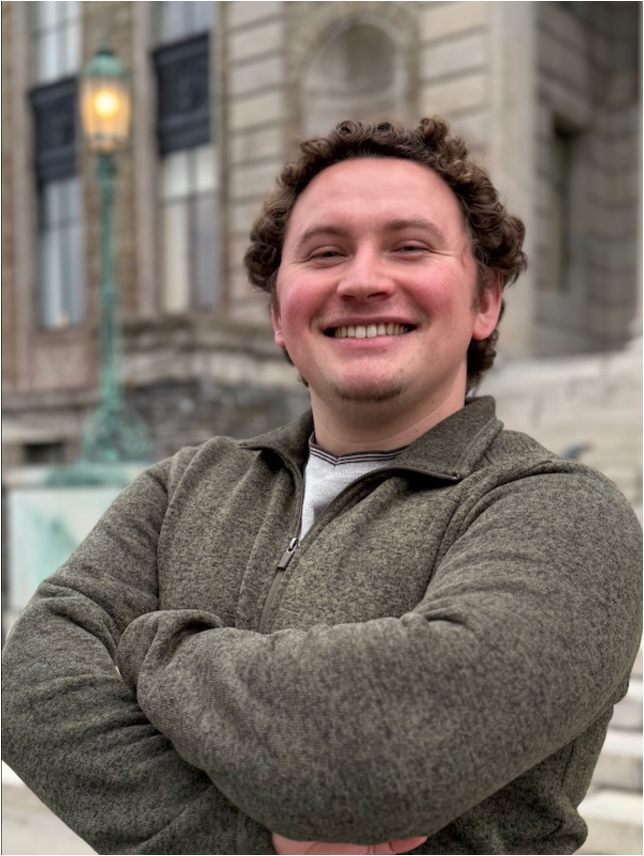 Smiling man with curly hair stands outdoors with arms crossed