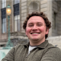 Smiling man with curly hair stands outdoors with arms crossed