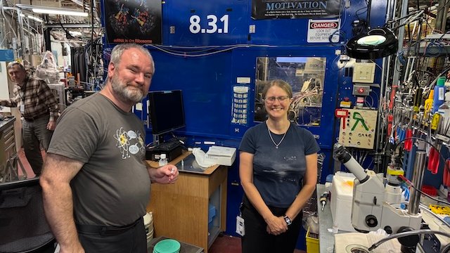 Two scientists stand beside laboratory equipment in a research facility