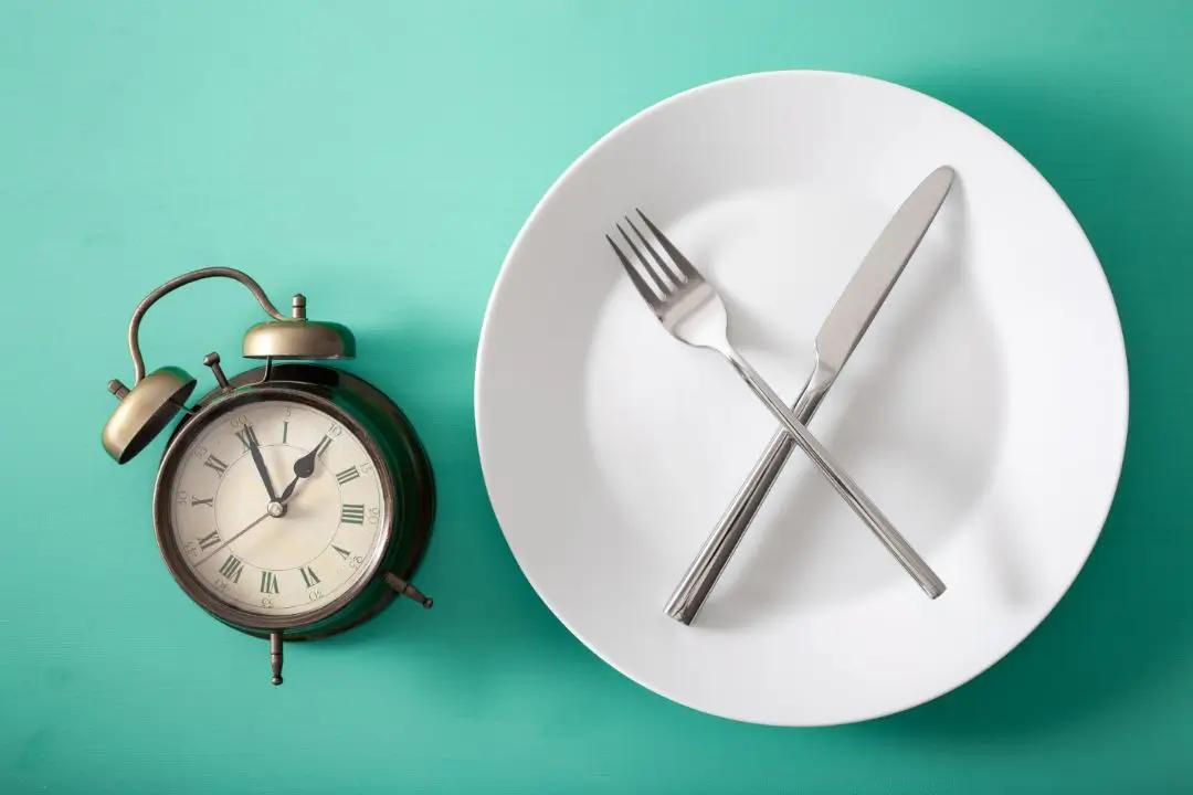An antique alarm clock sits next to a white plate with a fork and knife crossed in the center, all placed on a turquoise background.