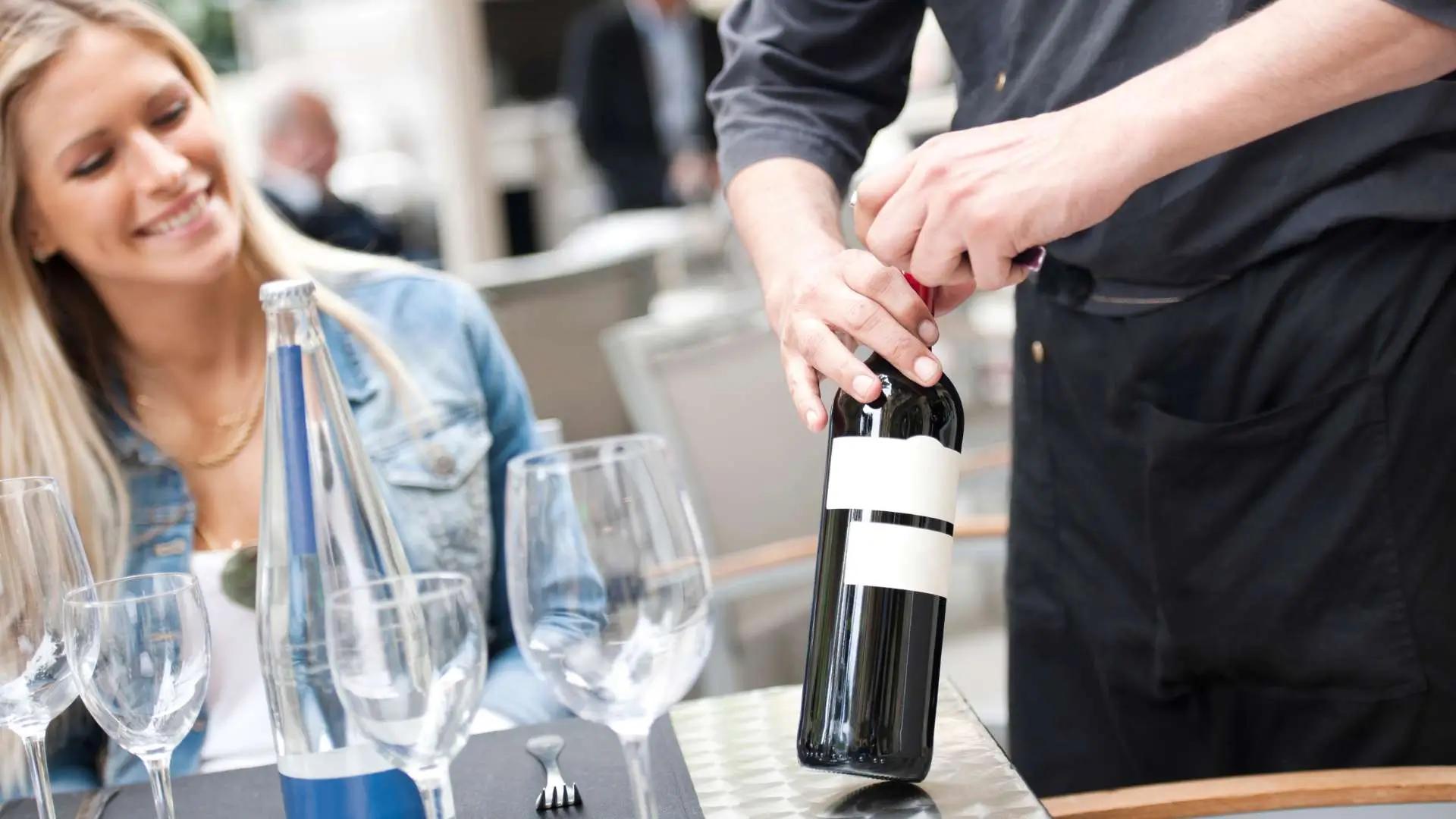 A server opening a bottle of wine at a table set with empty wine glasses and a bottle of water.