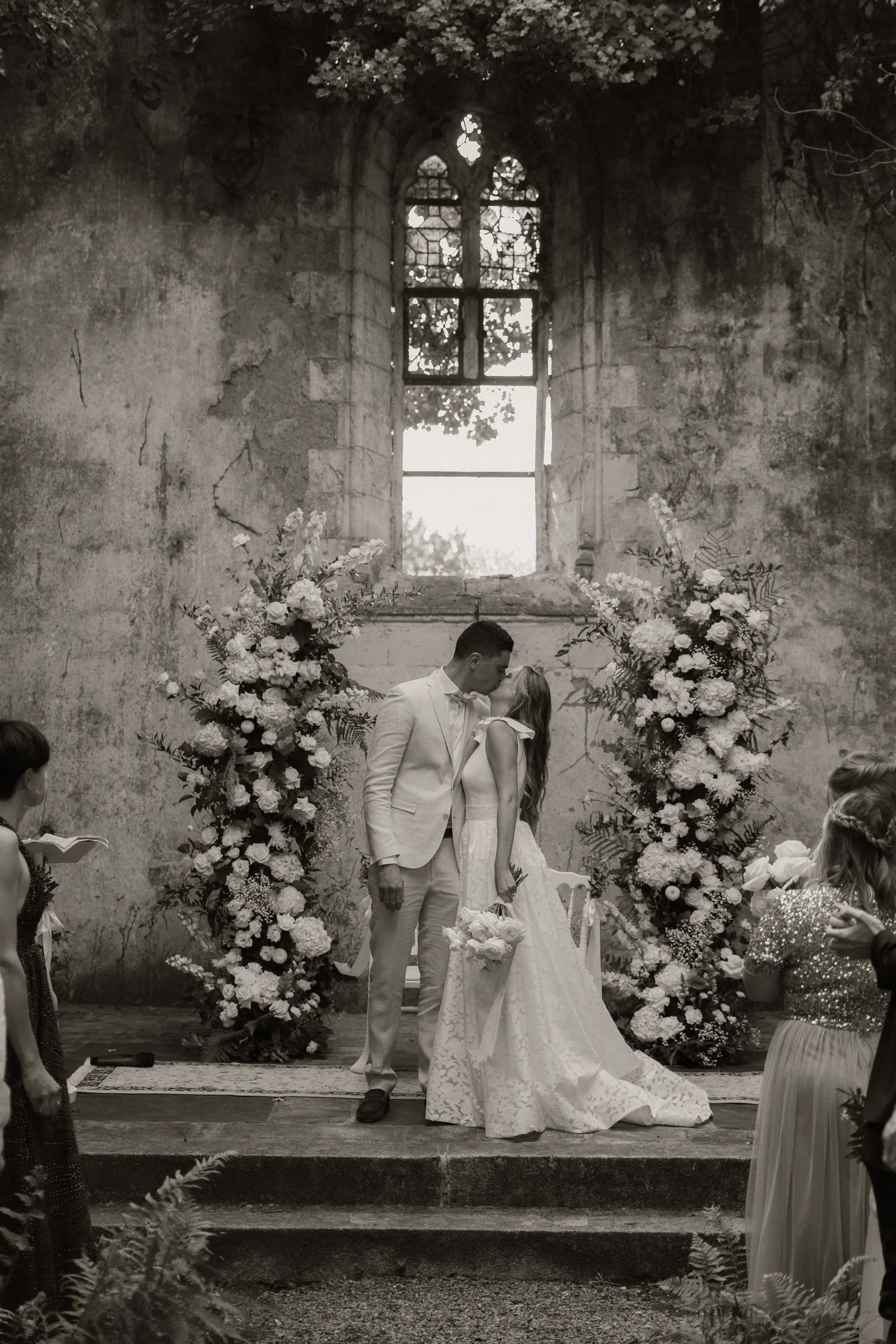 Baiser des mariés devant la fenêtre gothique de la chapelle, entre deux arches florales