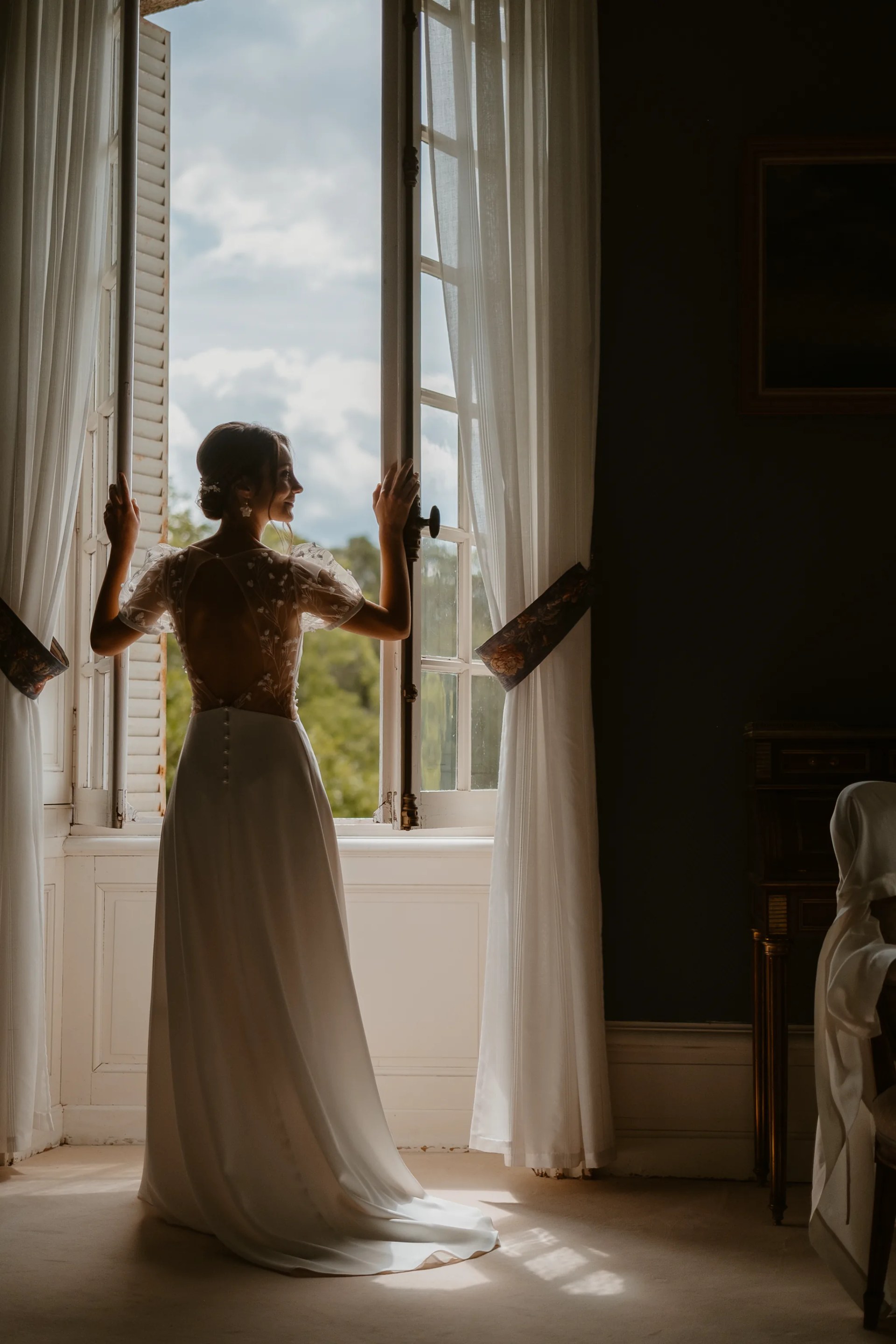 La mariée ouvrant les fenêtres de sa suite, robe de dentelle à dos ouvert