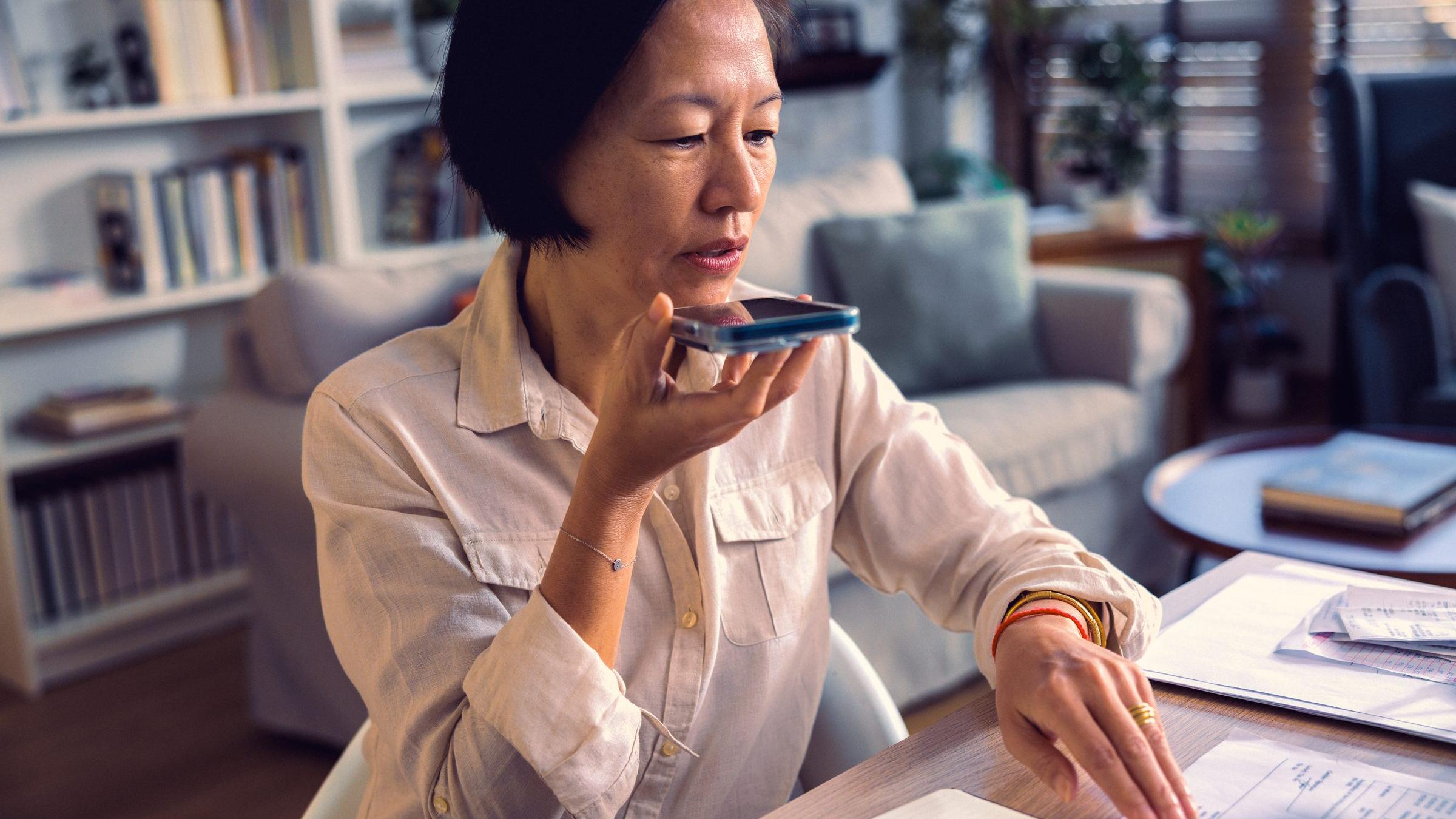 Woman at her desk at home speaking into her phone over paperwork