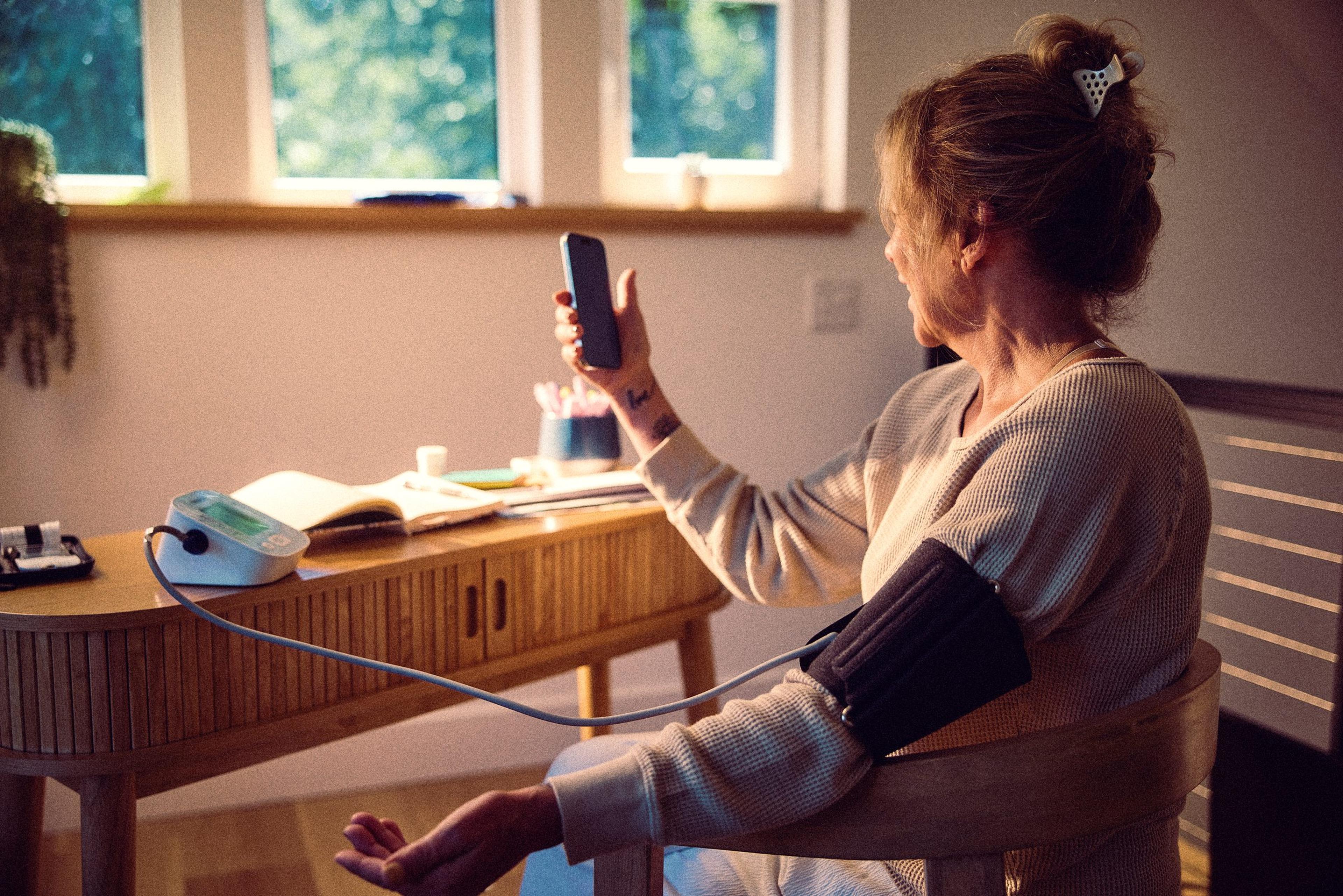 Woman at home holding up her phone while a blood pressure cuff takes a reading