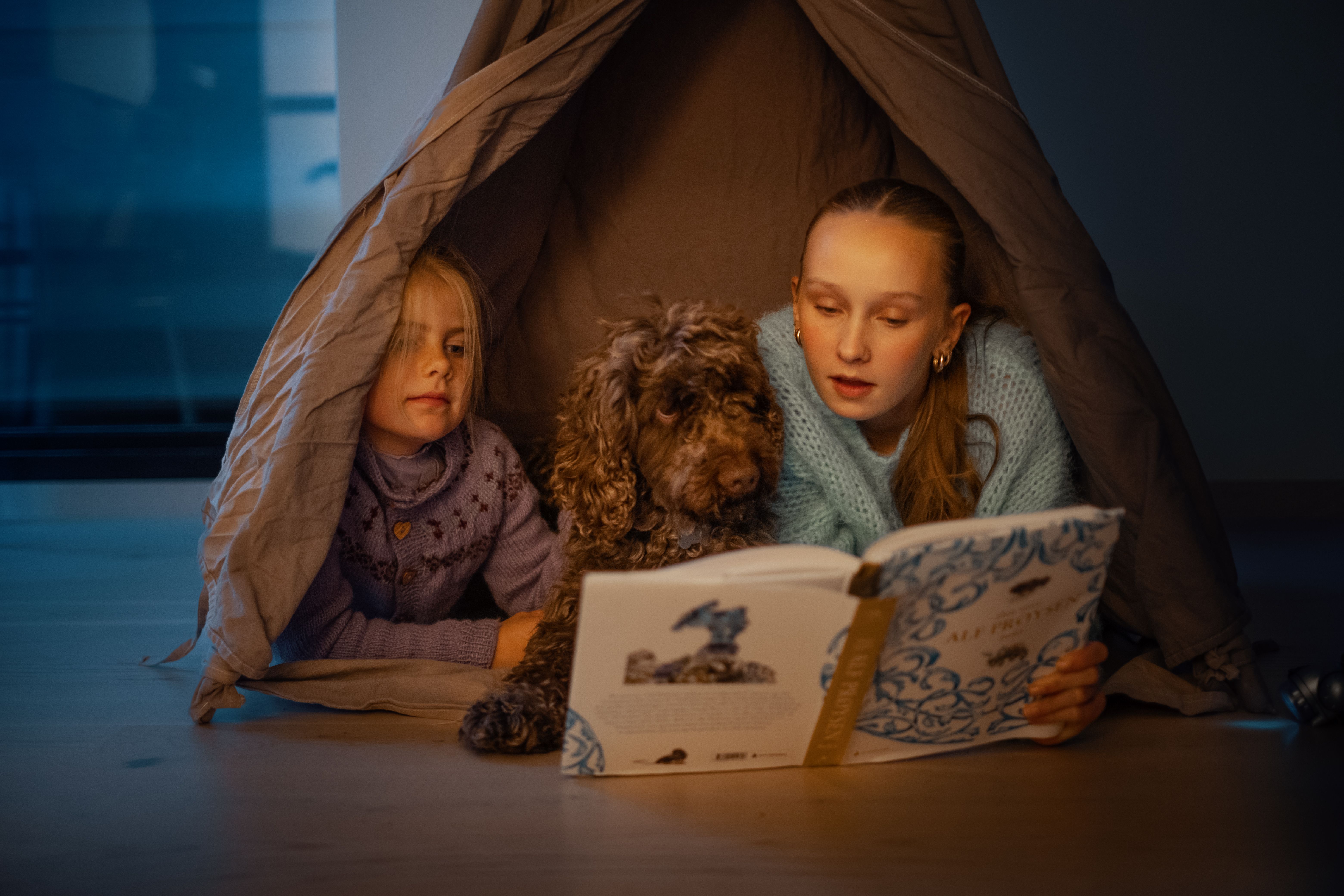 To jenter og en hund som leser bok delvis inne i et telt