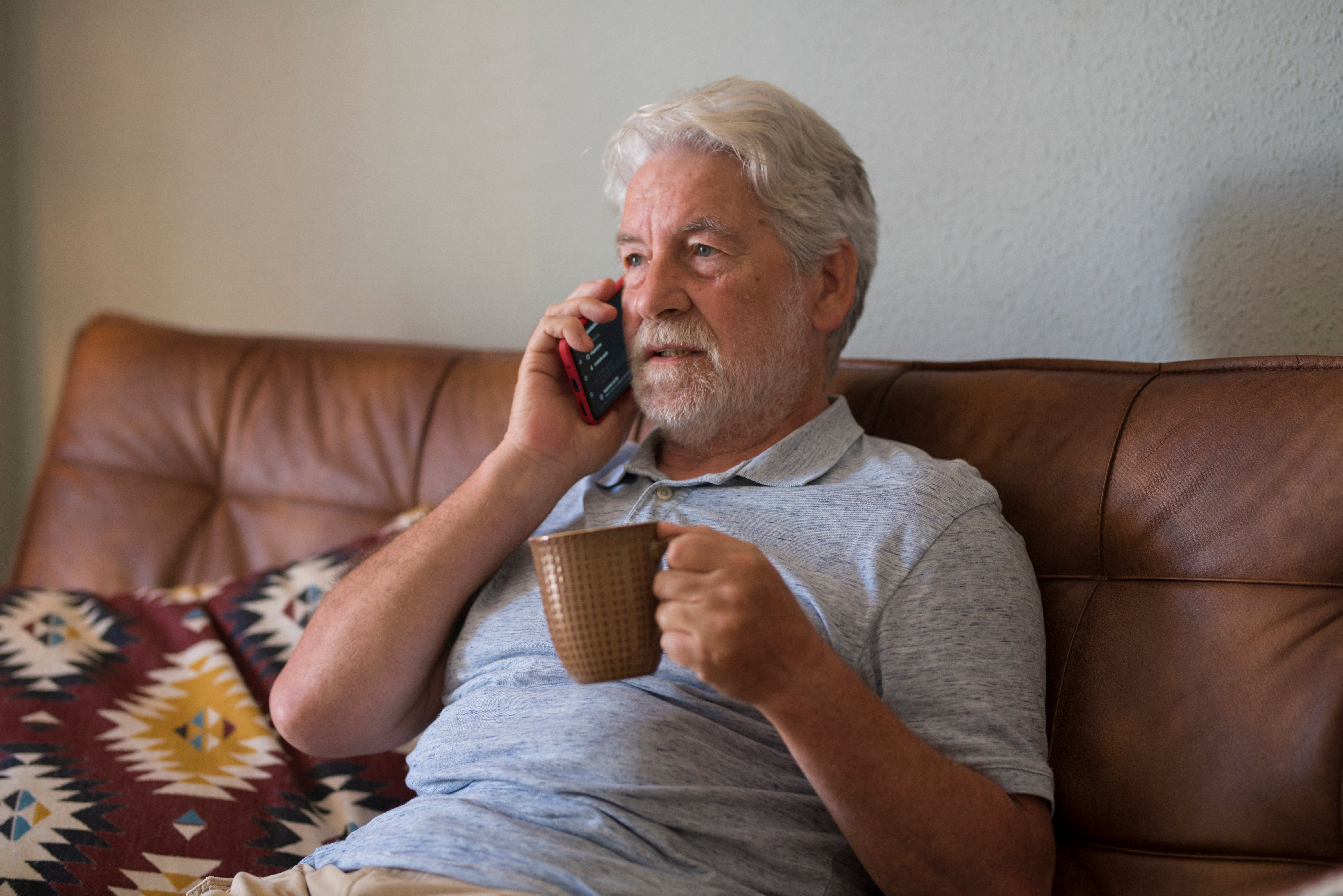 Older man sitting on couch with phone and hot drink 